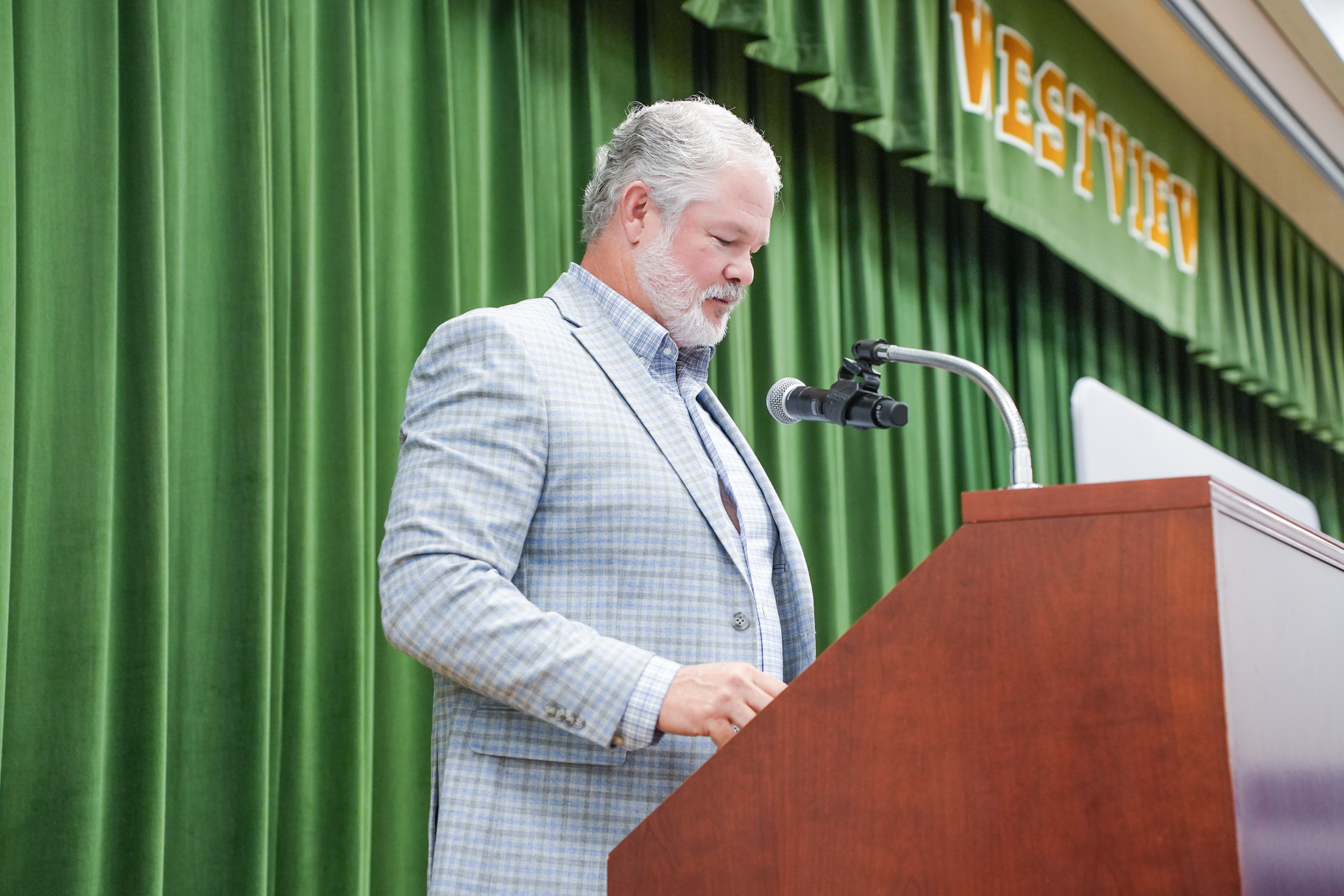 A man with white hair speaks at a podium with a microphone, wearing a light-colored suit. Green curtains are in the background.