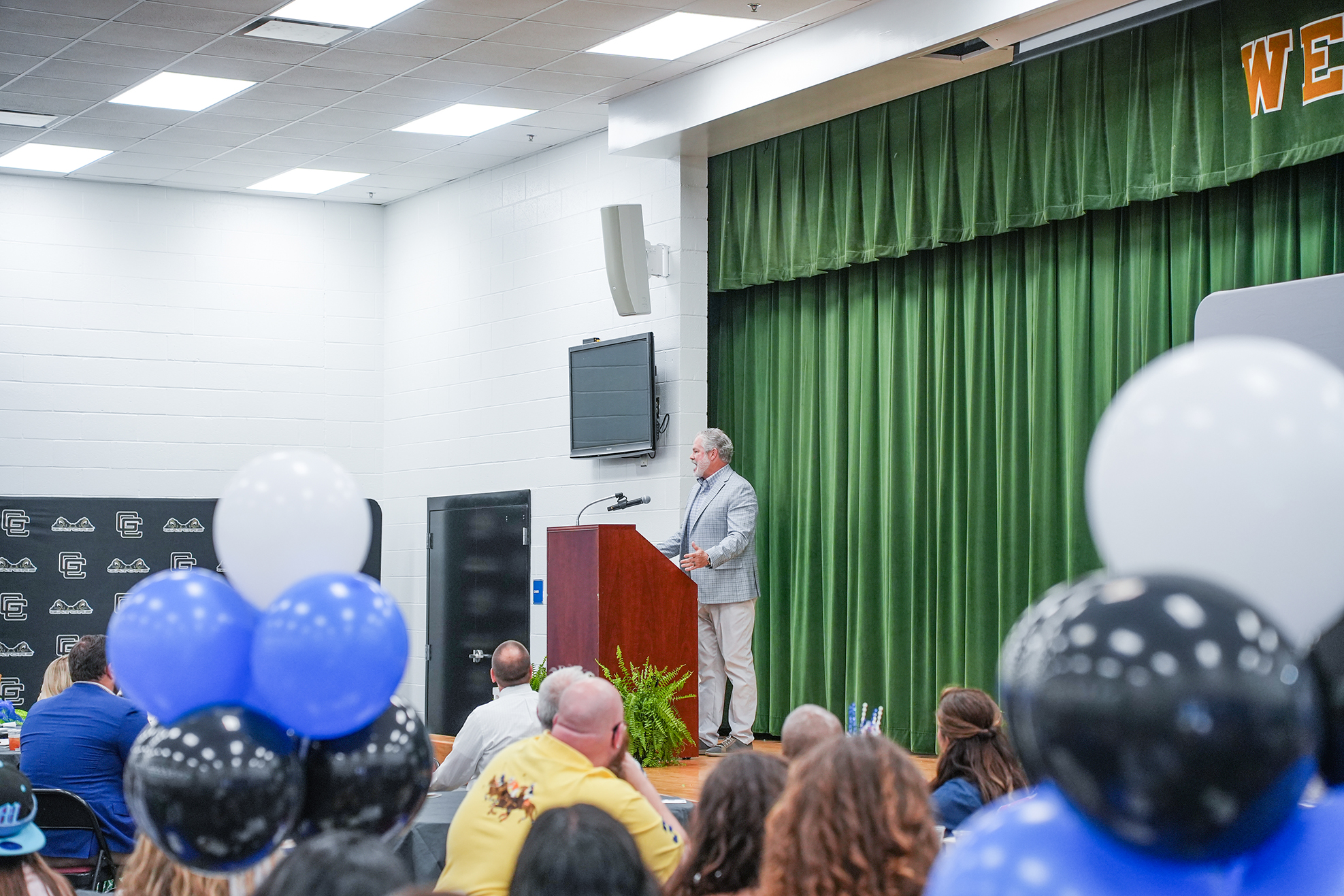 A man at a podium in a room with green curtains, a TV mounted on the wall, and a crowd of people.