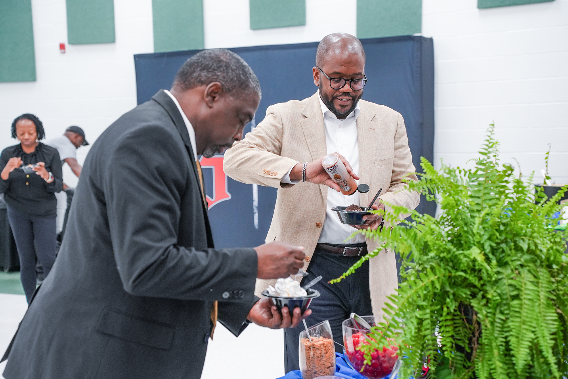 Two men in suits are at a table with food; one adds sauce to a bowl. A plant is nearby.