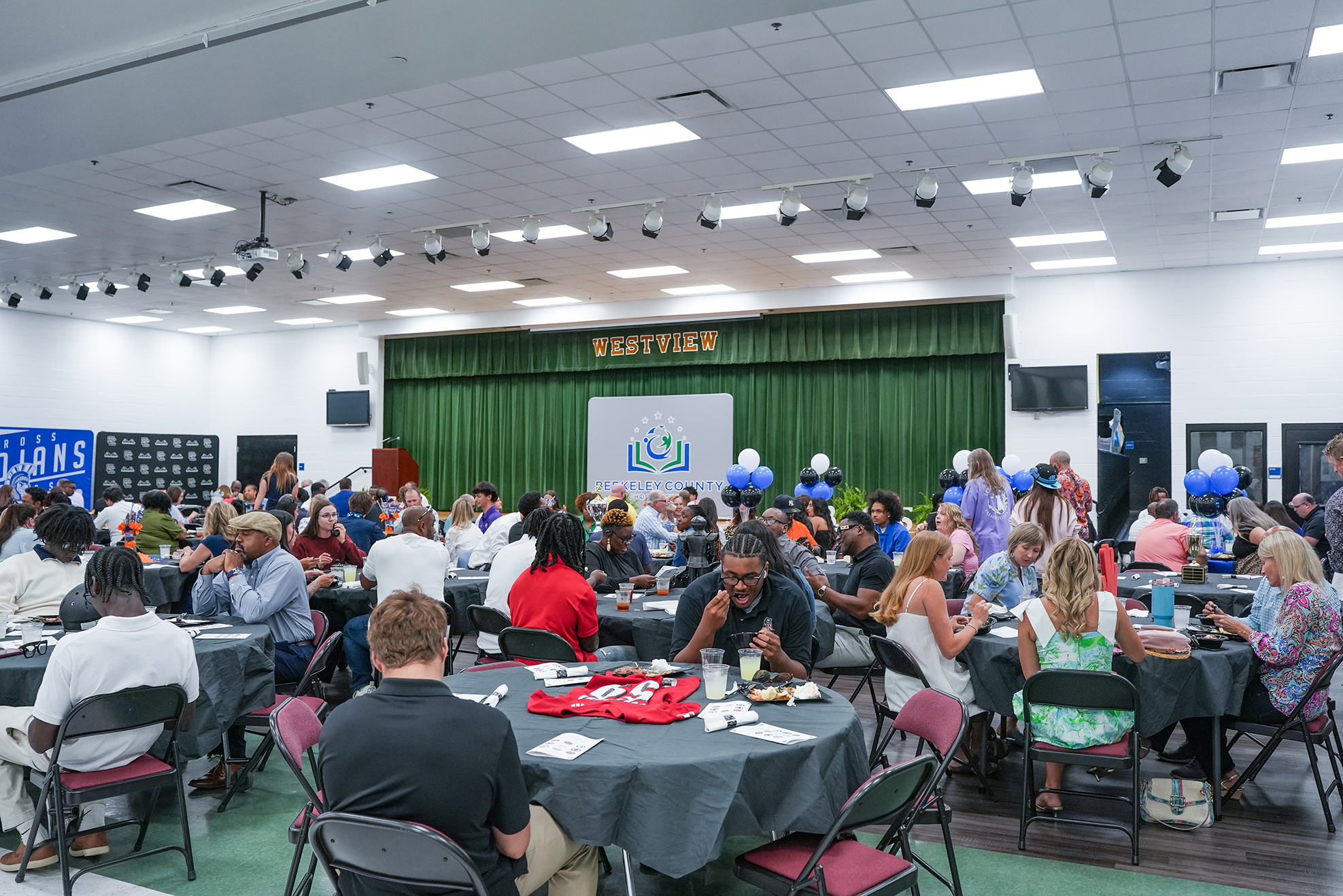 Indoor dining area with tables and chairs. A green curtain and banner behind. A crowd of people sitting and standing.