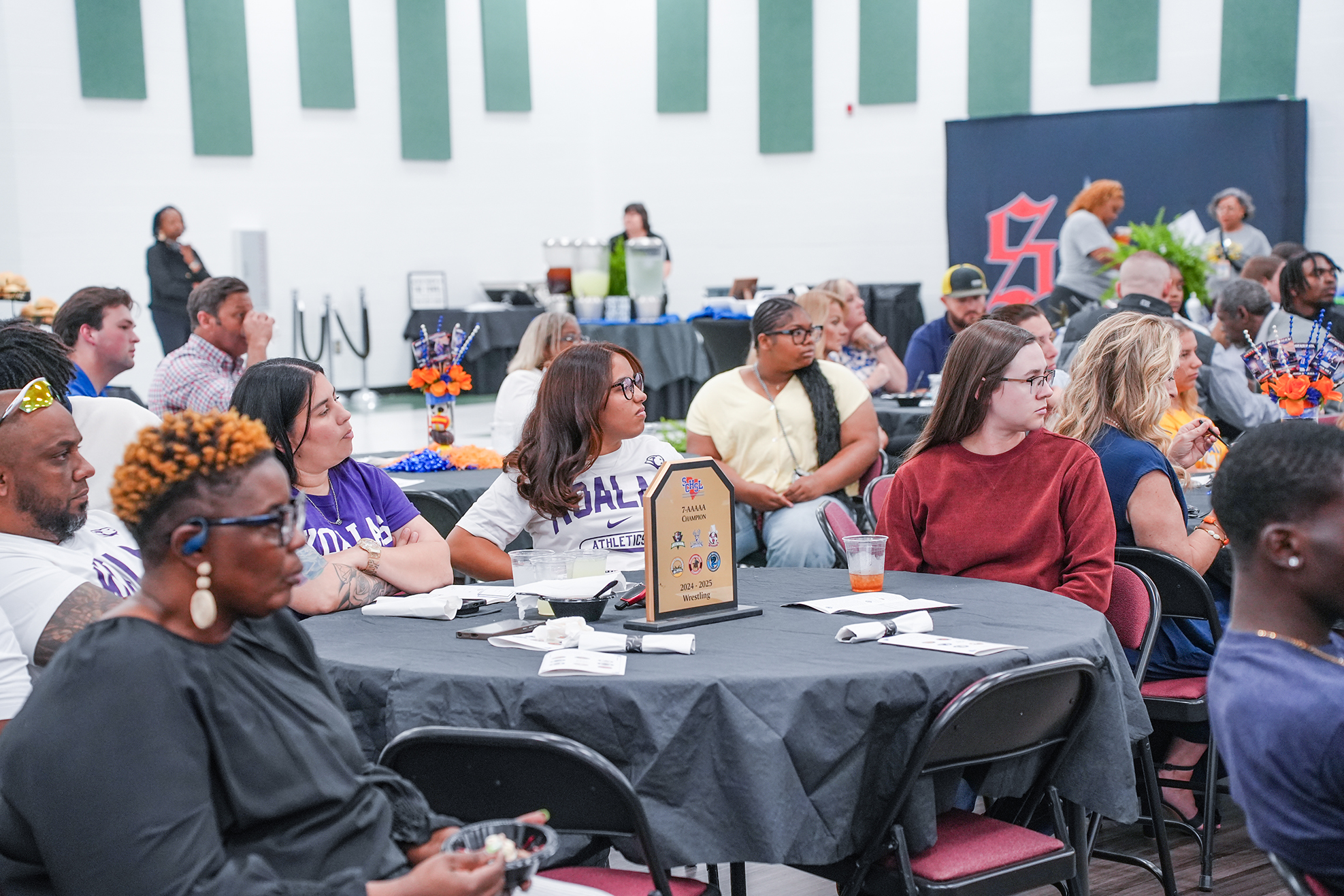 People seated at tables in an indoor space, with some standing by tables and a banner visible in the background.
