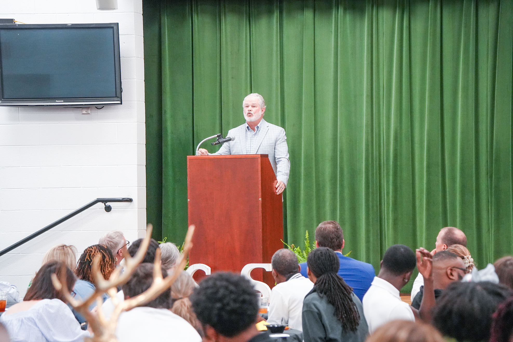 A man speaks at a podium in a room with a green curtain; several people sit in front of him.