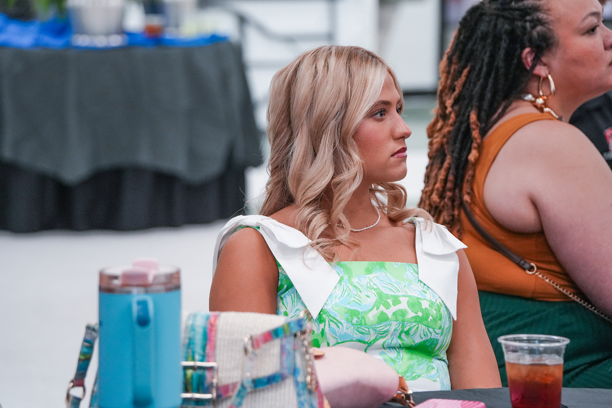 Two women sit at a table, one wearing a green dress, the other with braided hair. Nearby, a table holds various items.