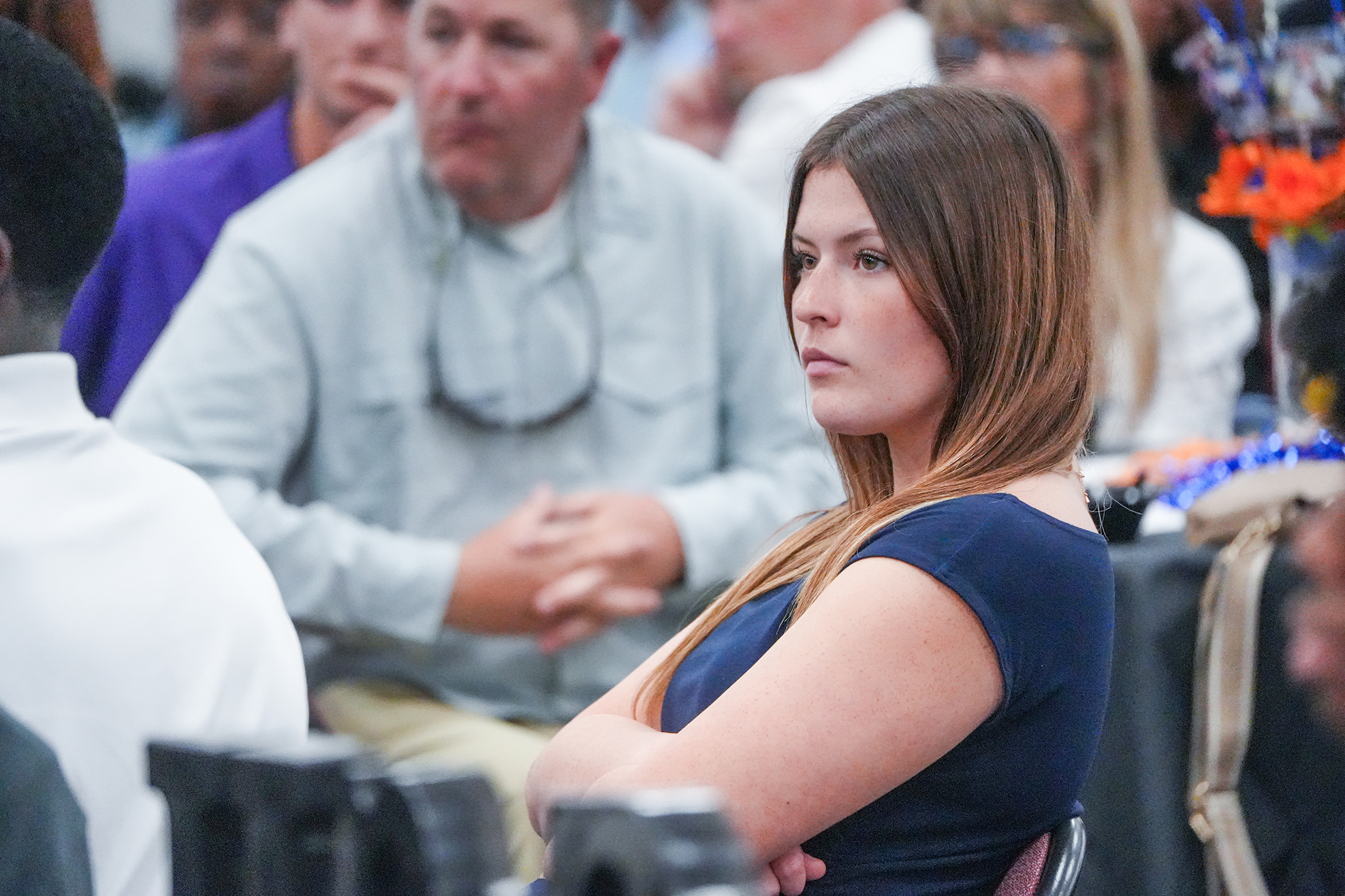 A woman in a blue top sits with her arms crossed, facing a crowd of people.