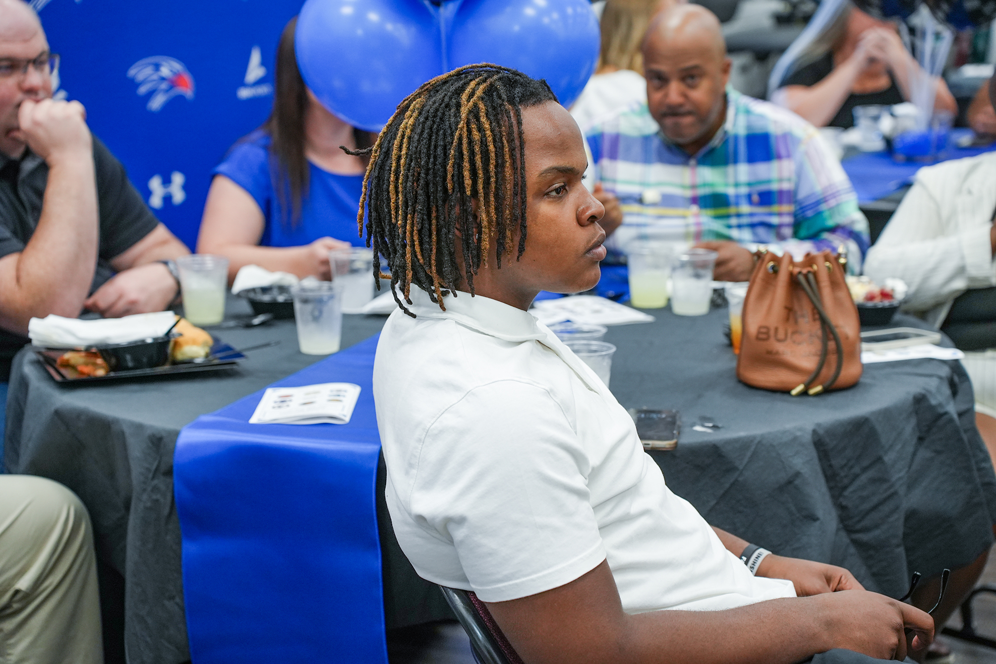 A person with braided hair sits at a table with others; some tables have balloons and handbags.