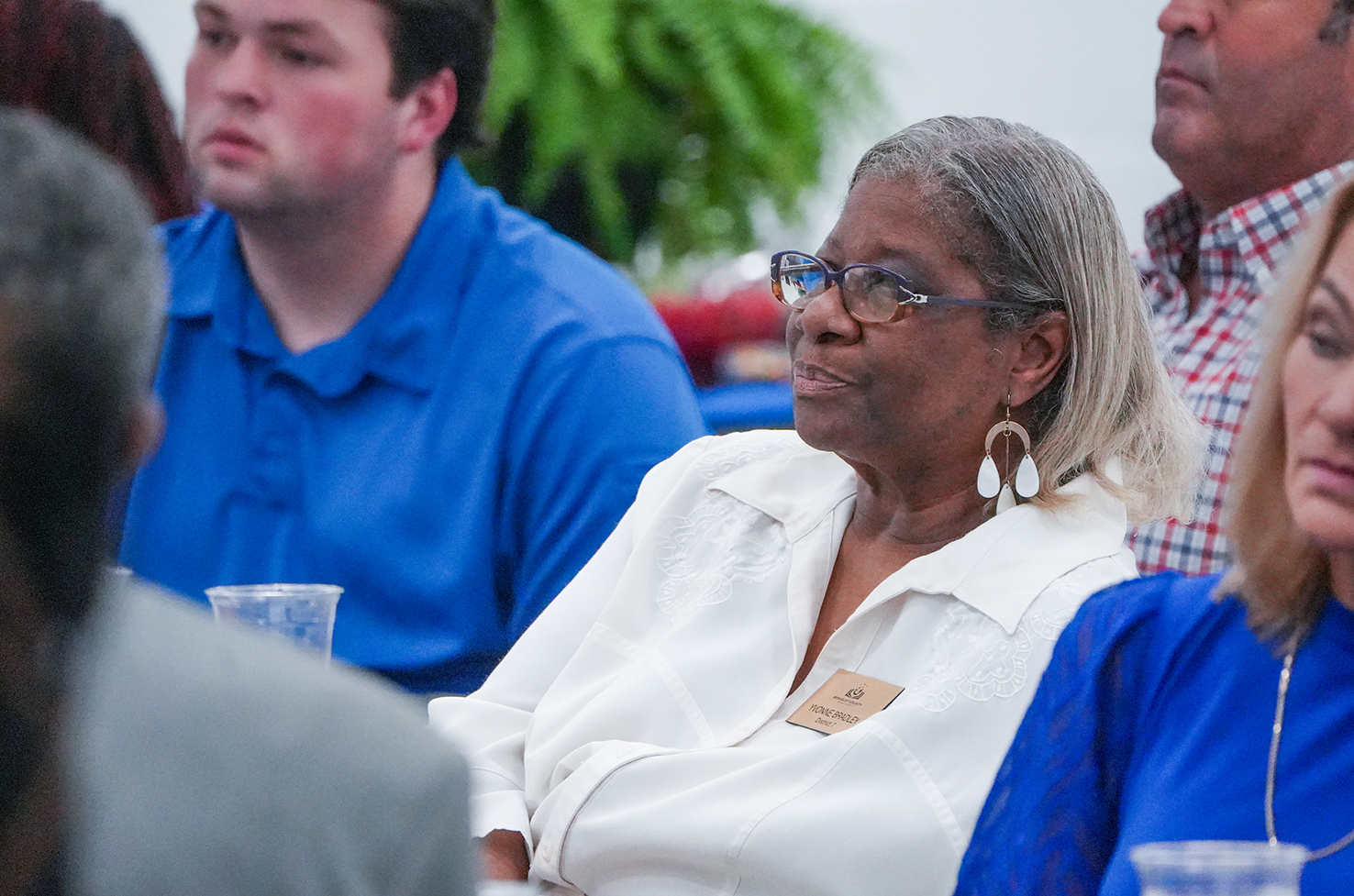 Several adults in a conference room with a man in a blue shirt, an older woman in a white blouse, and a woman in a blue shirt.