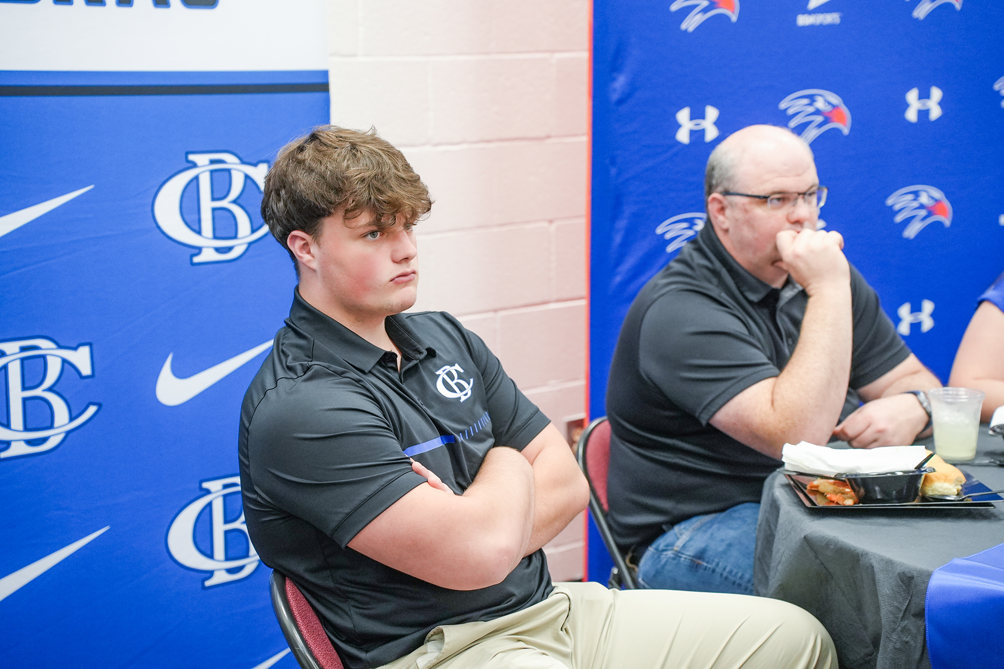 Two individuals seated at a table, one with arms crossed, other resting chin on hand. Background includes a blue banner.