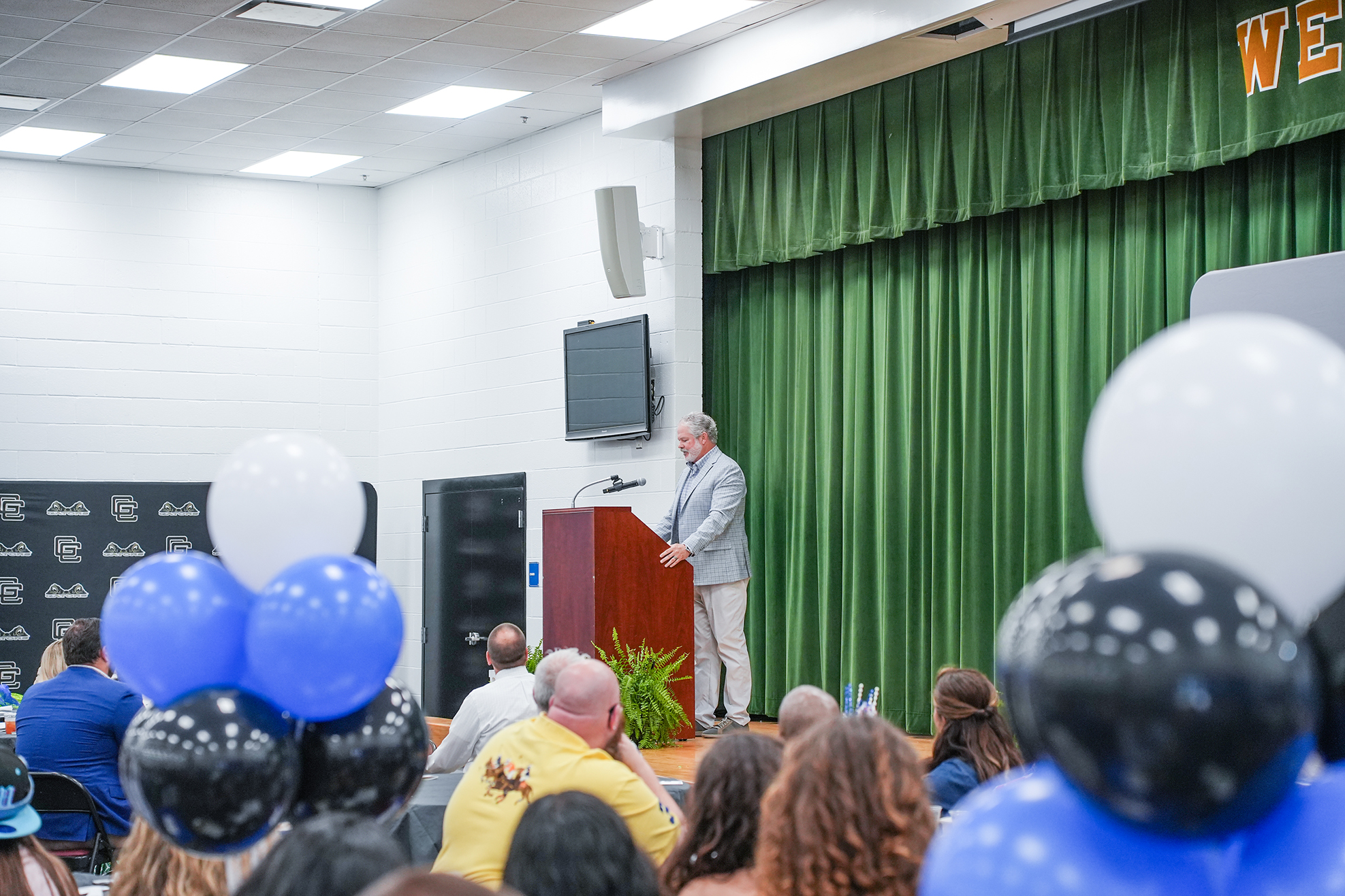 A person stands at a podium with a microphone in a room filled with people. Green curtains are in the background.