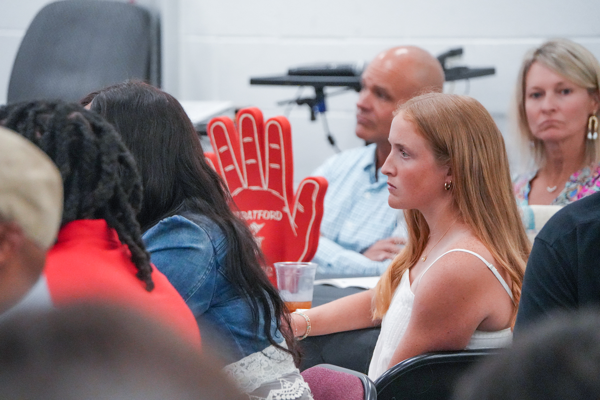 Group of people in a classroom or meeting room, some wearing jewelry. One holds a red glove.
