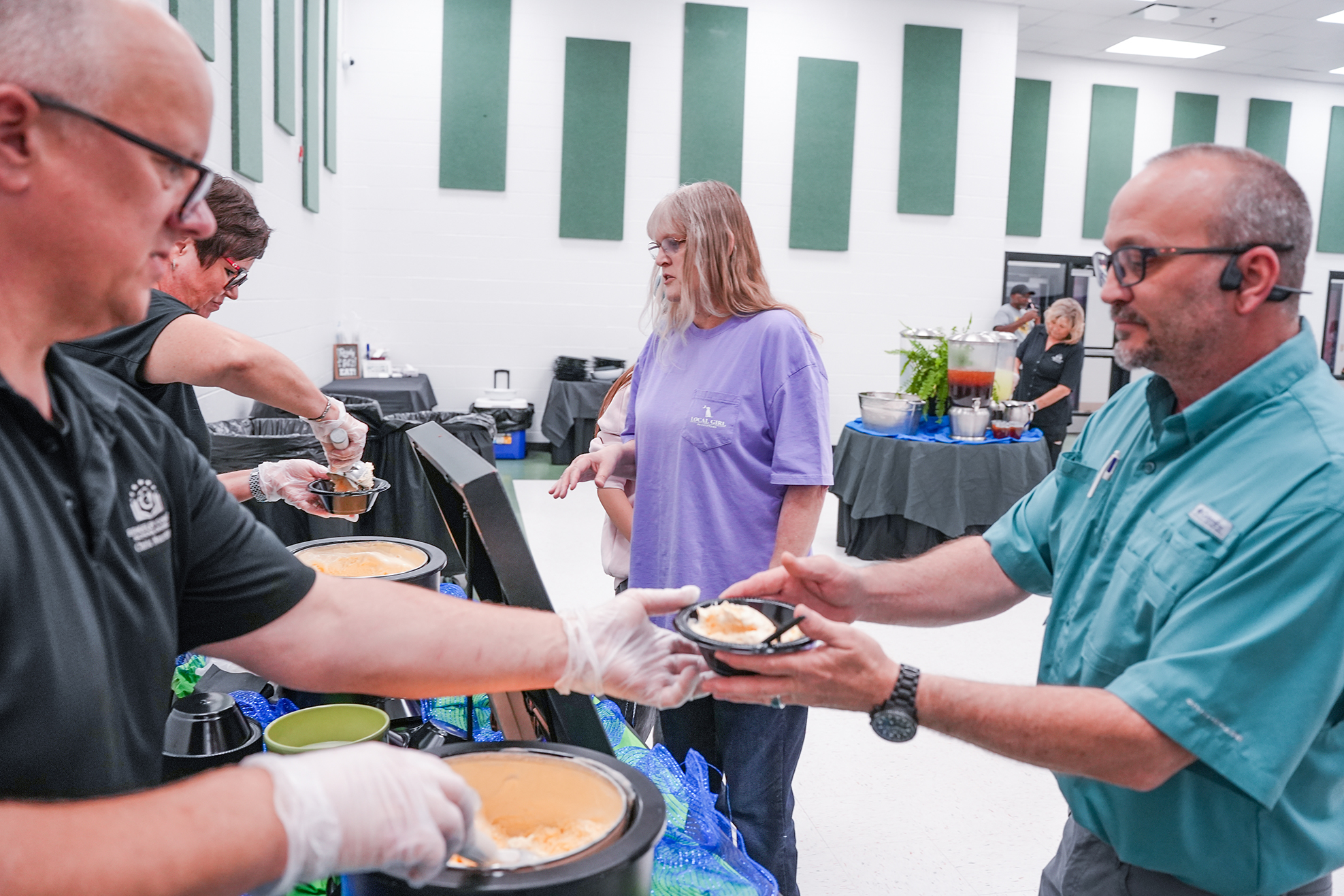 People in a cafeteria prepare food; one hands a plate to another. Background includes tables, plants, and a green-paneled wall.