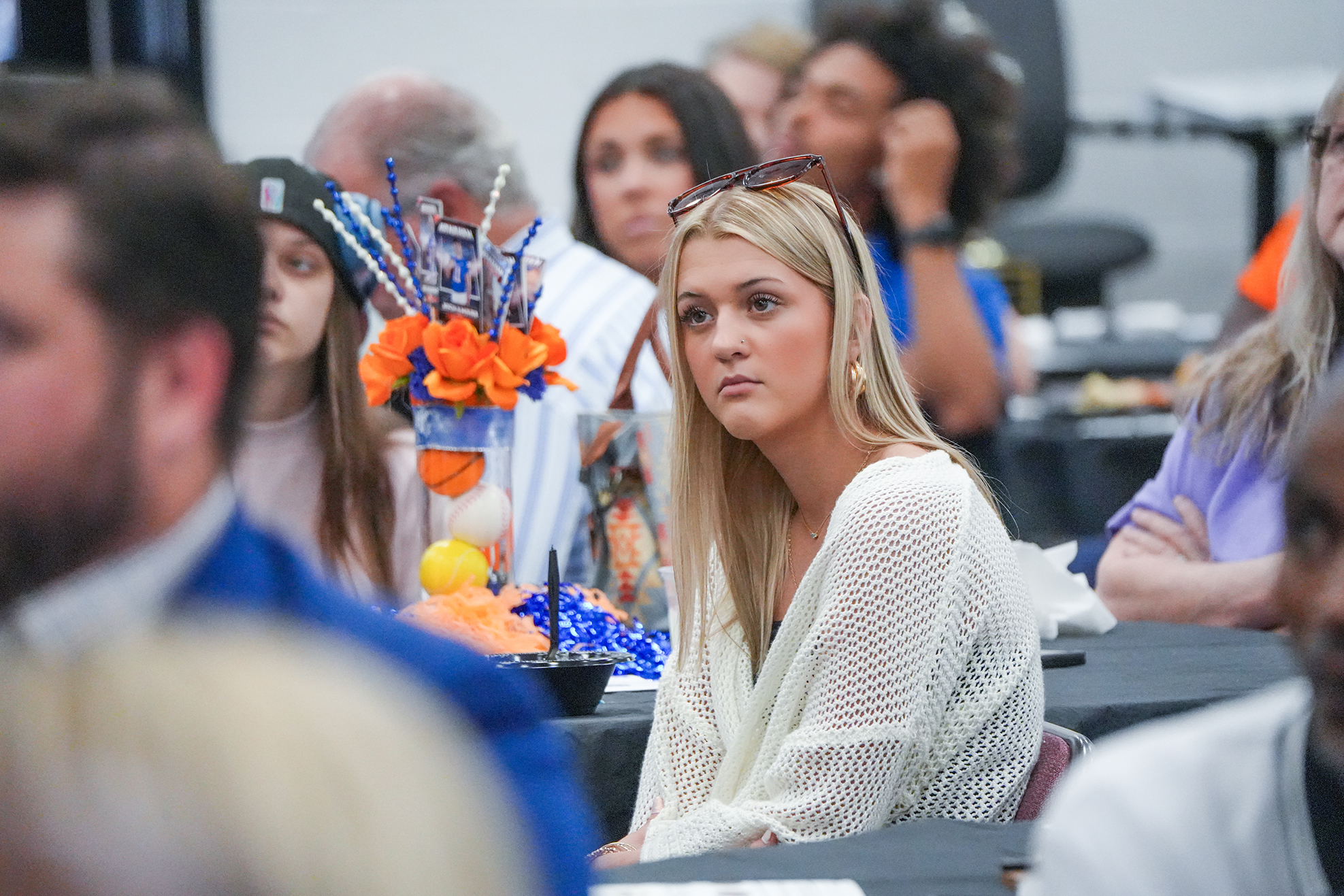 People sit around a table. A woman in white looks away from a floral centerpiece.