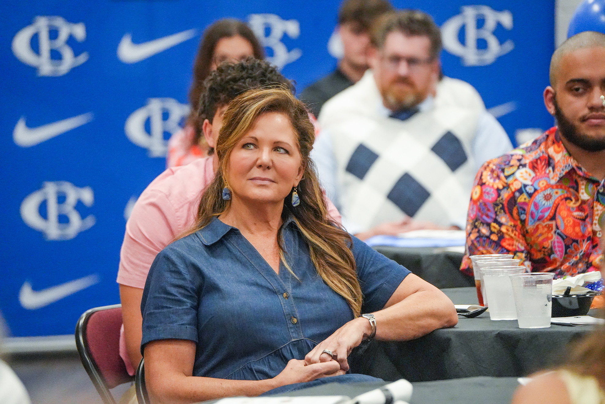 A woman in a blue shirt sits at a table with her arms crossed, surrounded by others.
