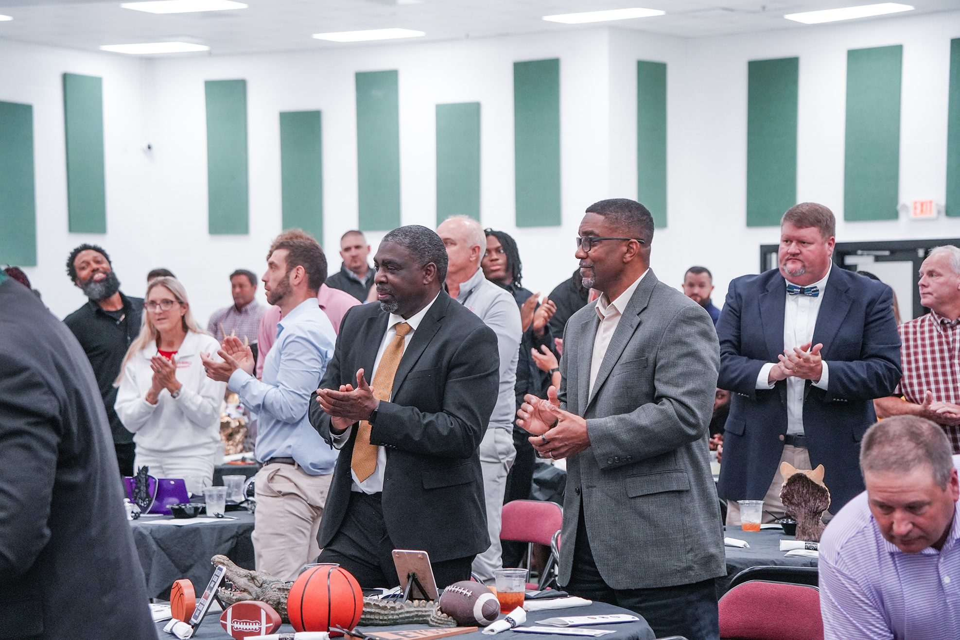A group of people in business attire are clapping inside a room with tables and sports items.