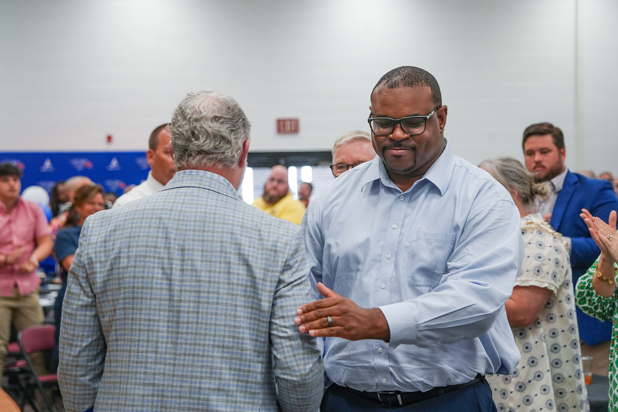 Indoor room with people standing and talking. Man in blue shirt and glasses talking to another man.