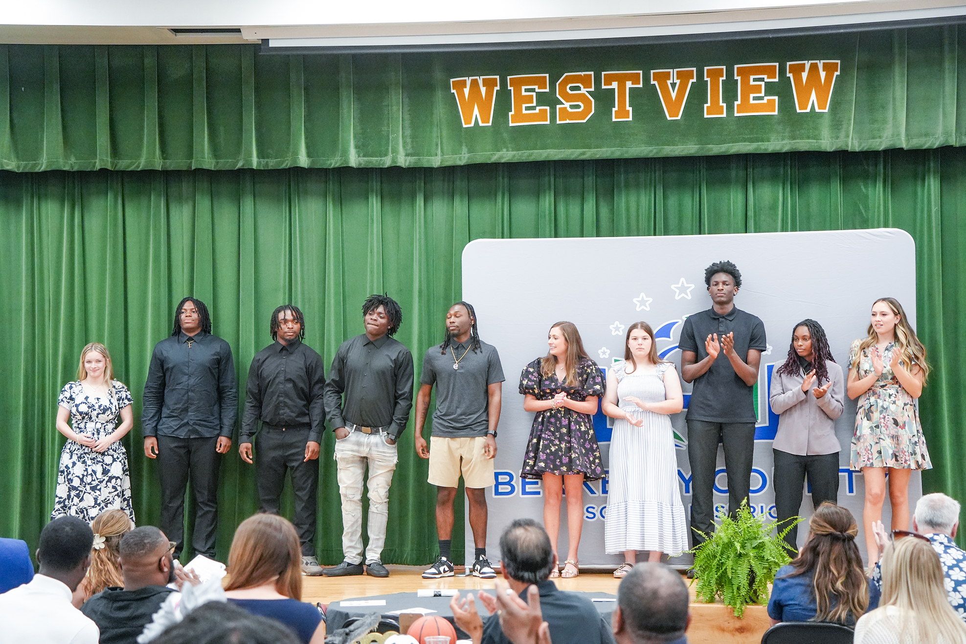 A group of students and faculty stands on a stage with "Westview" behind them, clapping and smiling.