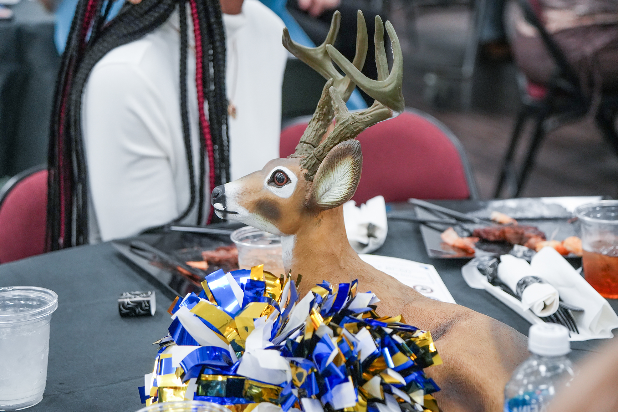 A table holds a faux deer, a bottle, a cup, and a pom-pom. A person sits in the background.