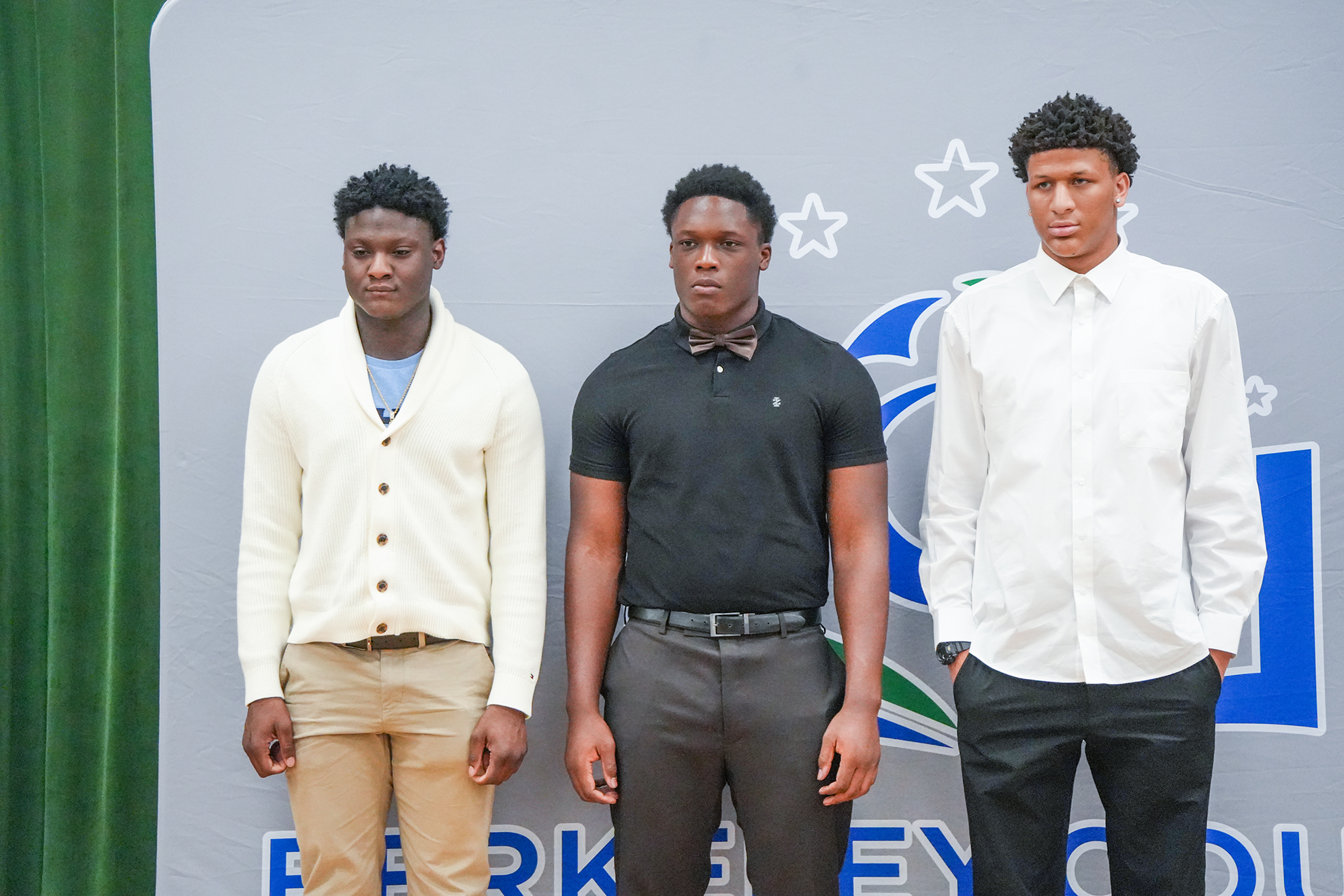 Three young men stand next to each other on a stage, wearing formal attire. A gray backdrop has a logo and stars.