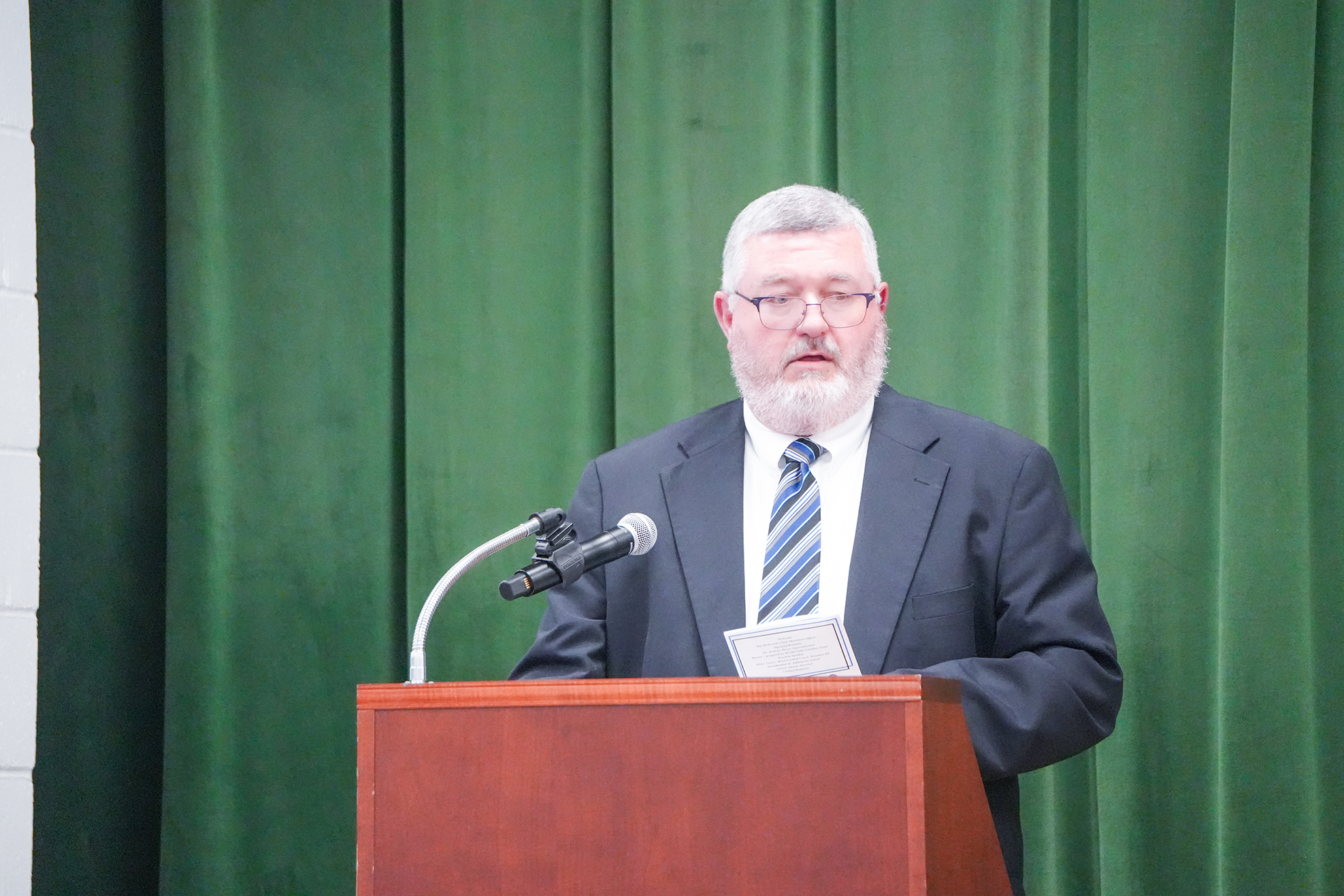 Man in suit and glasses at a podium, speaking into a microphone. Behind him is a green curtain.