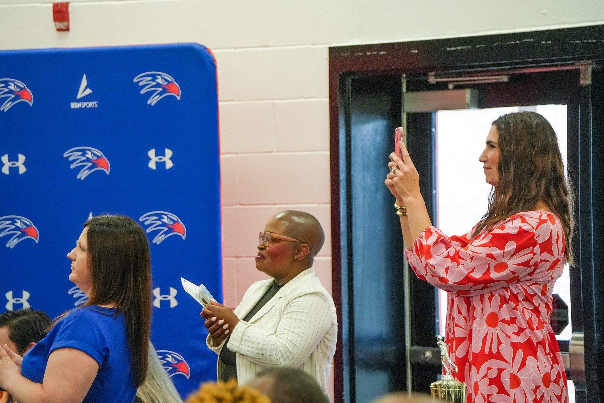 Three women face a blue backdrop featuring a logo. The woman in white speaks to the woman in blue.