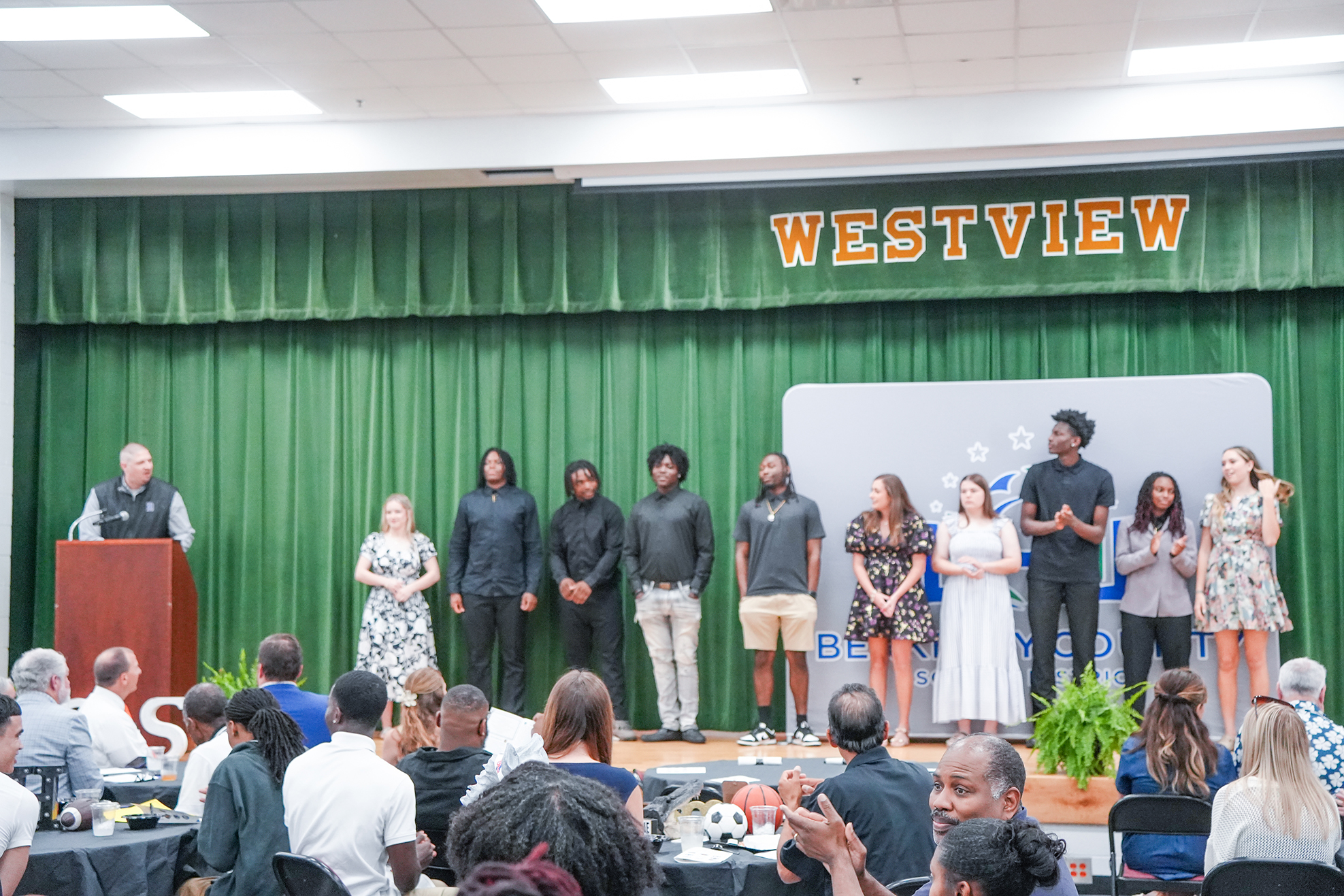 People stand on a stage, facing an audience, with a green curtain behind them and a banner reading "WESTVIEW."