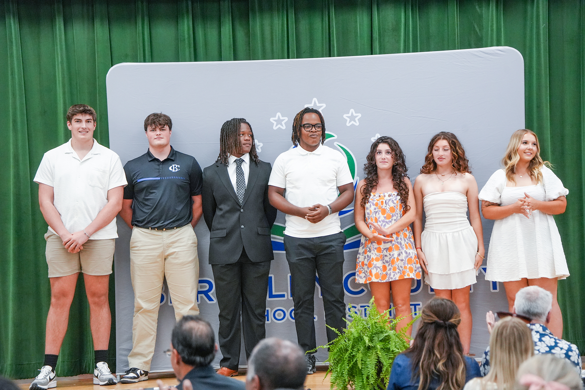 Five men and four women on stage in front of a green backdrop and audience.