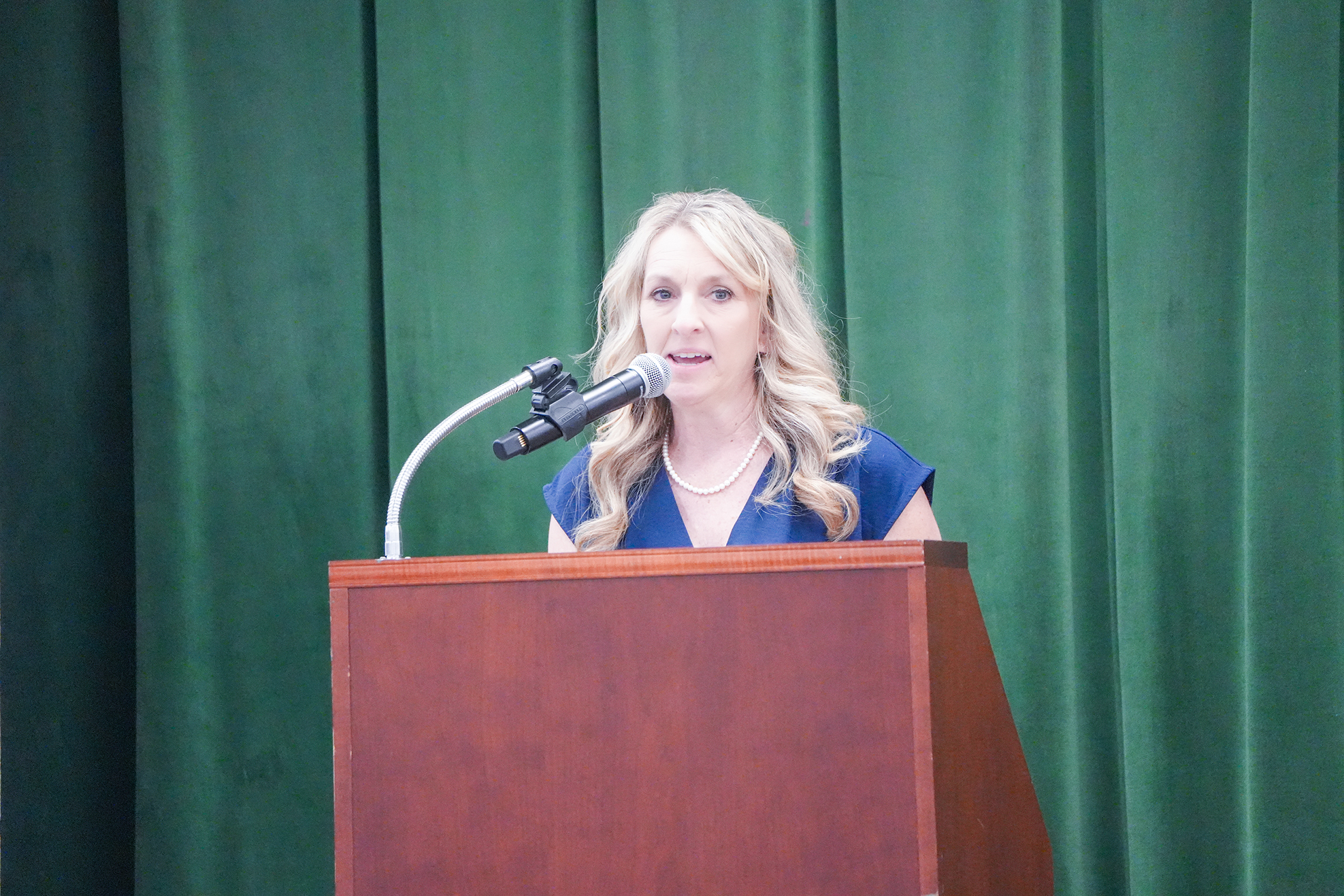 A woman with blonde hair stands at a podium with a microphone, wearing a blue top. Behind her, a green curtain.