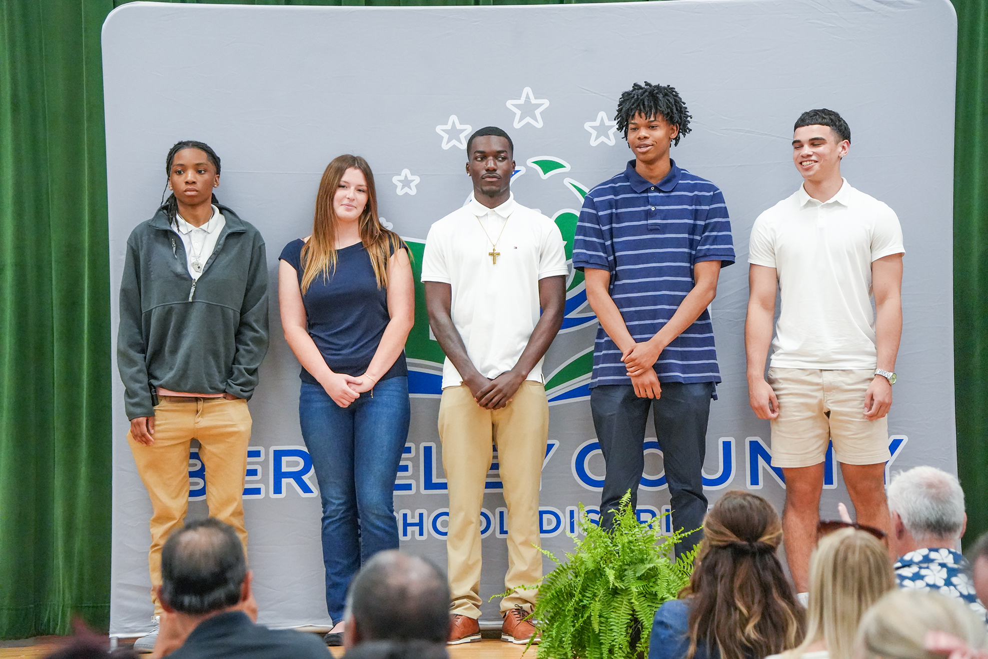 Five individuals stand on a stage, facing the audience. They wear casual clothes. Behind them is a backdrop with text and symbols.