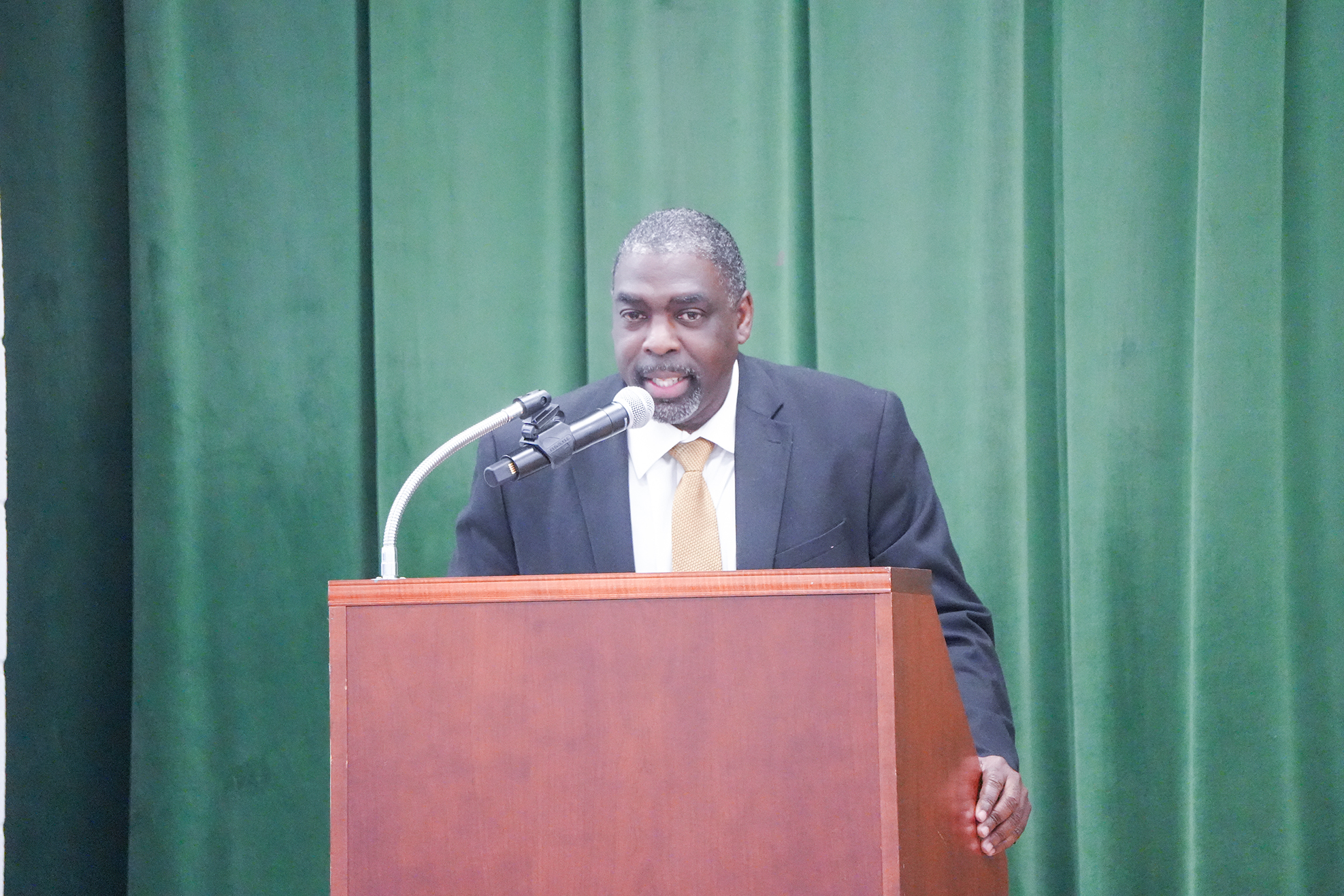 A man in a suit and tie stands at a podium with a microphone against a green curtain.