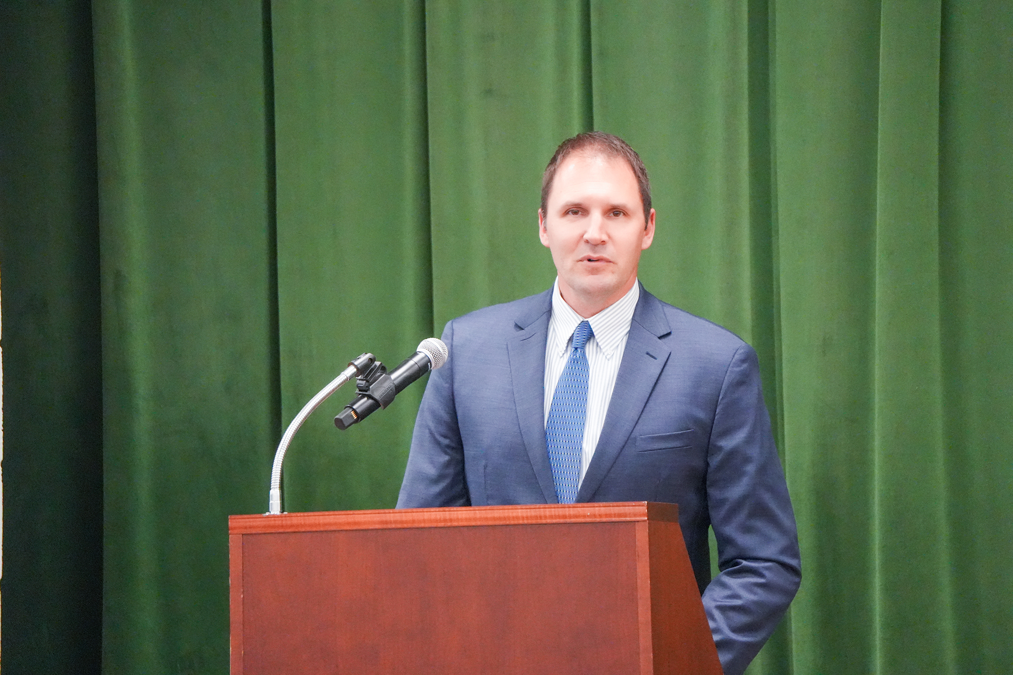 A man in a business suit and tie stands behind a podium, addressing an audience with a microphone.