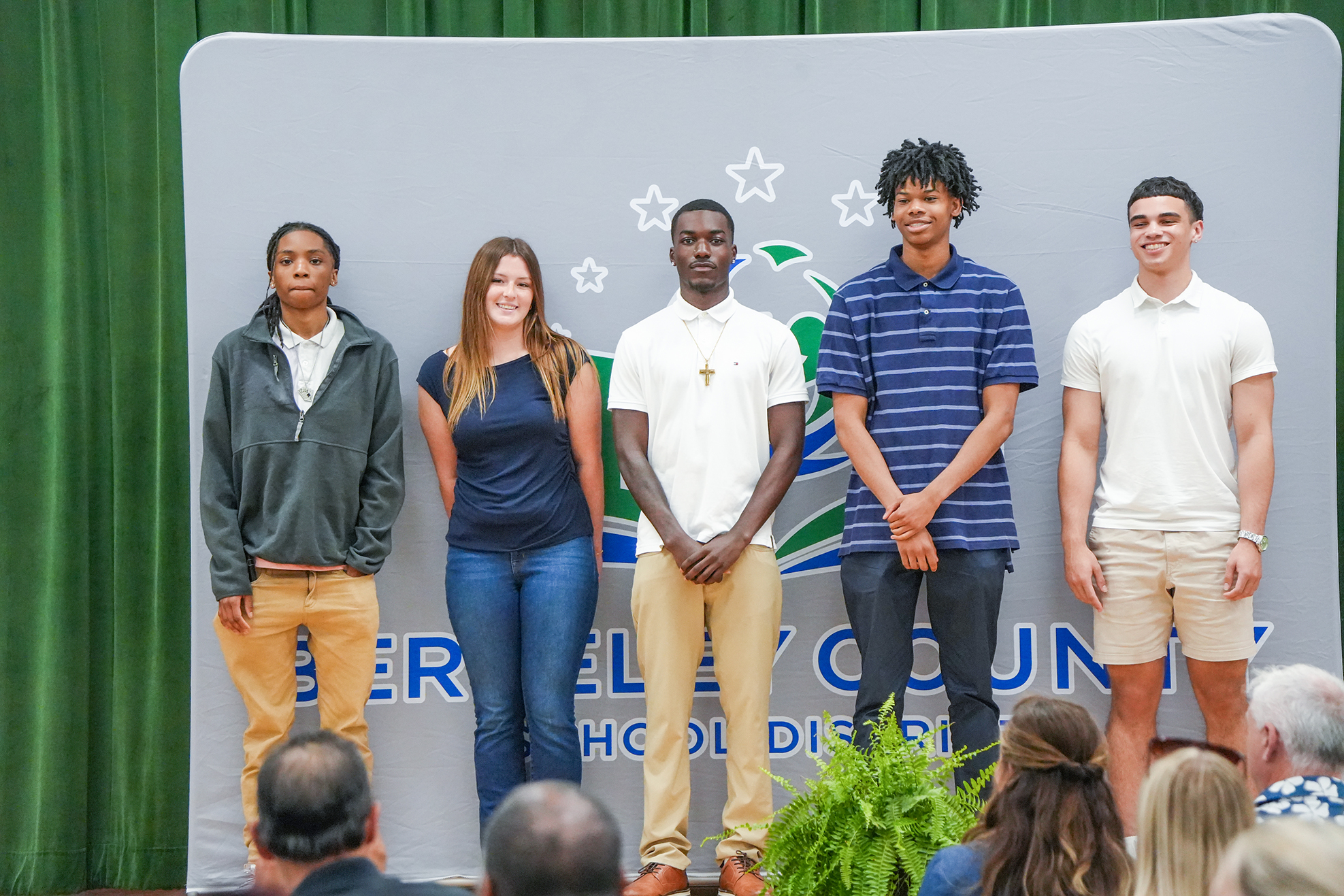 Five young adults stand on a stage with a backdrop. They are smiling and appear to be waiting for a ceremony to start.