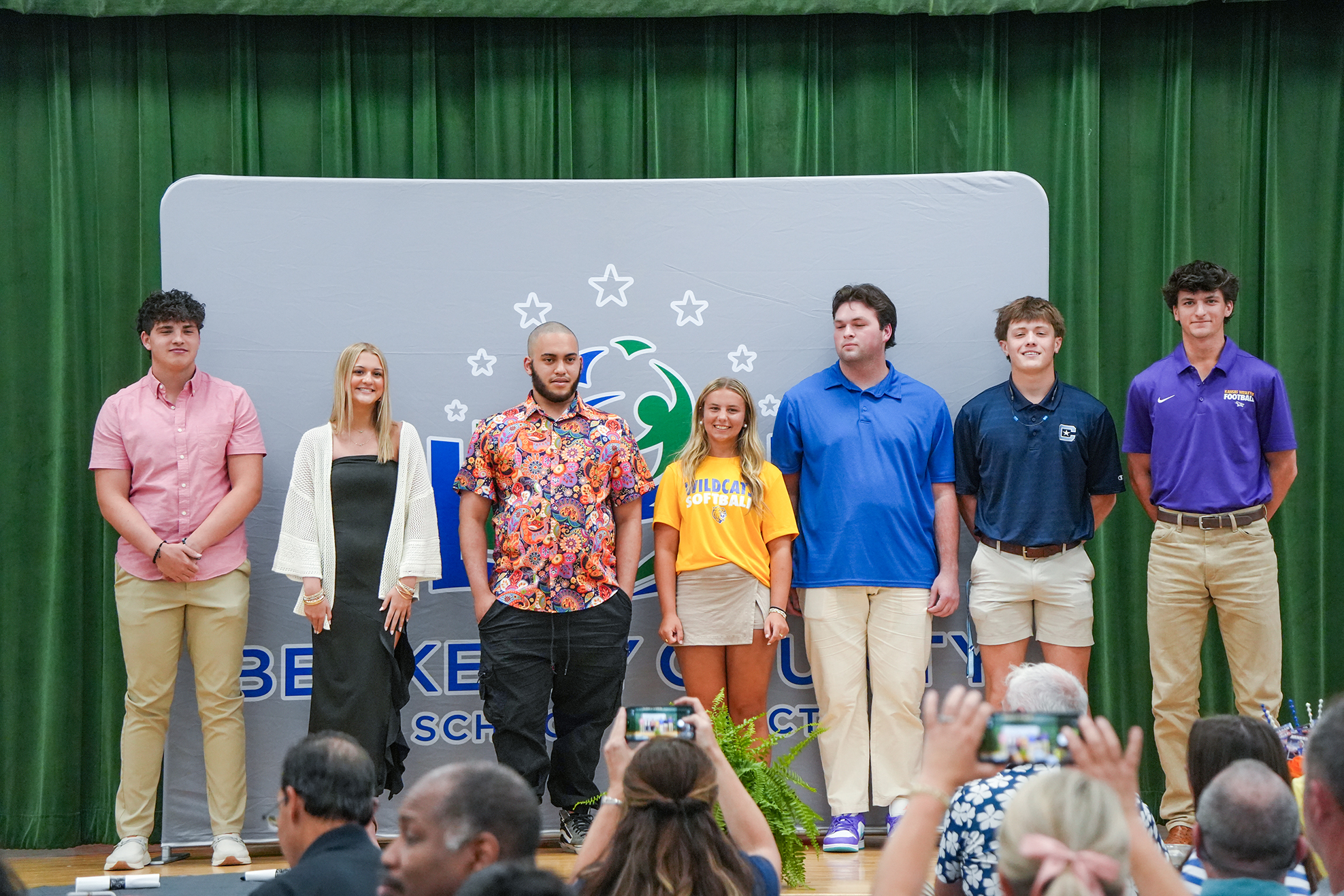 Six people on a stage, likely at an awards ceremony. Behind them, a backdrop displays "BE" and "SCHOOL" with logos. Audience members seated in front.