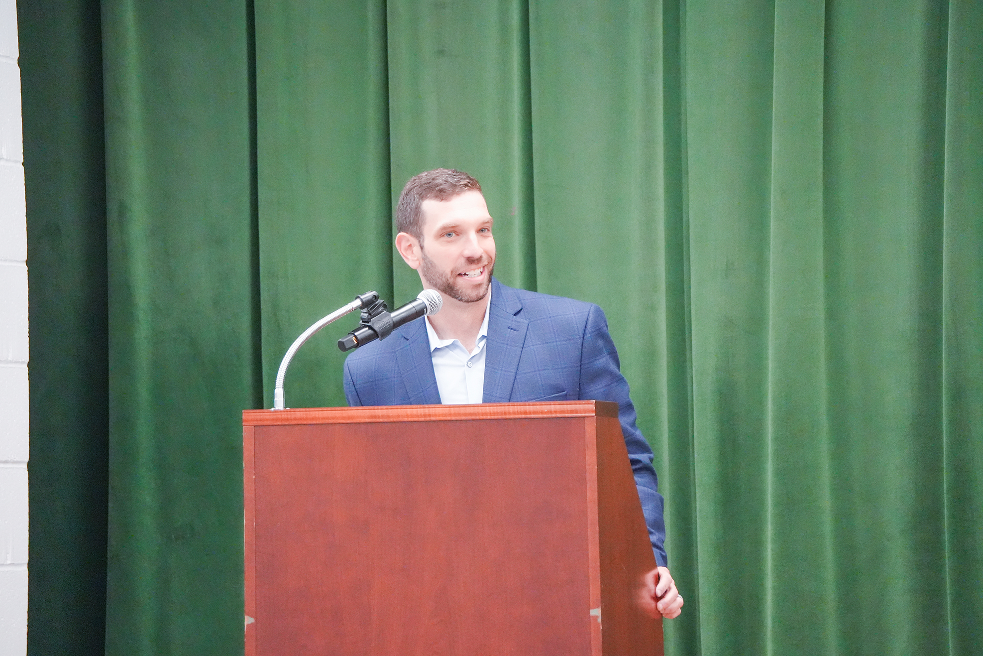 A man speaks into a microphone at a podium with a green curtain in the background.