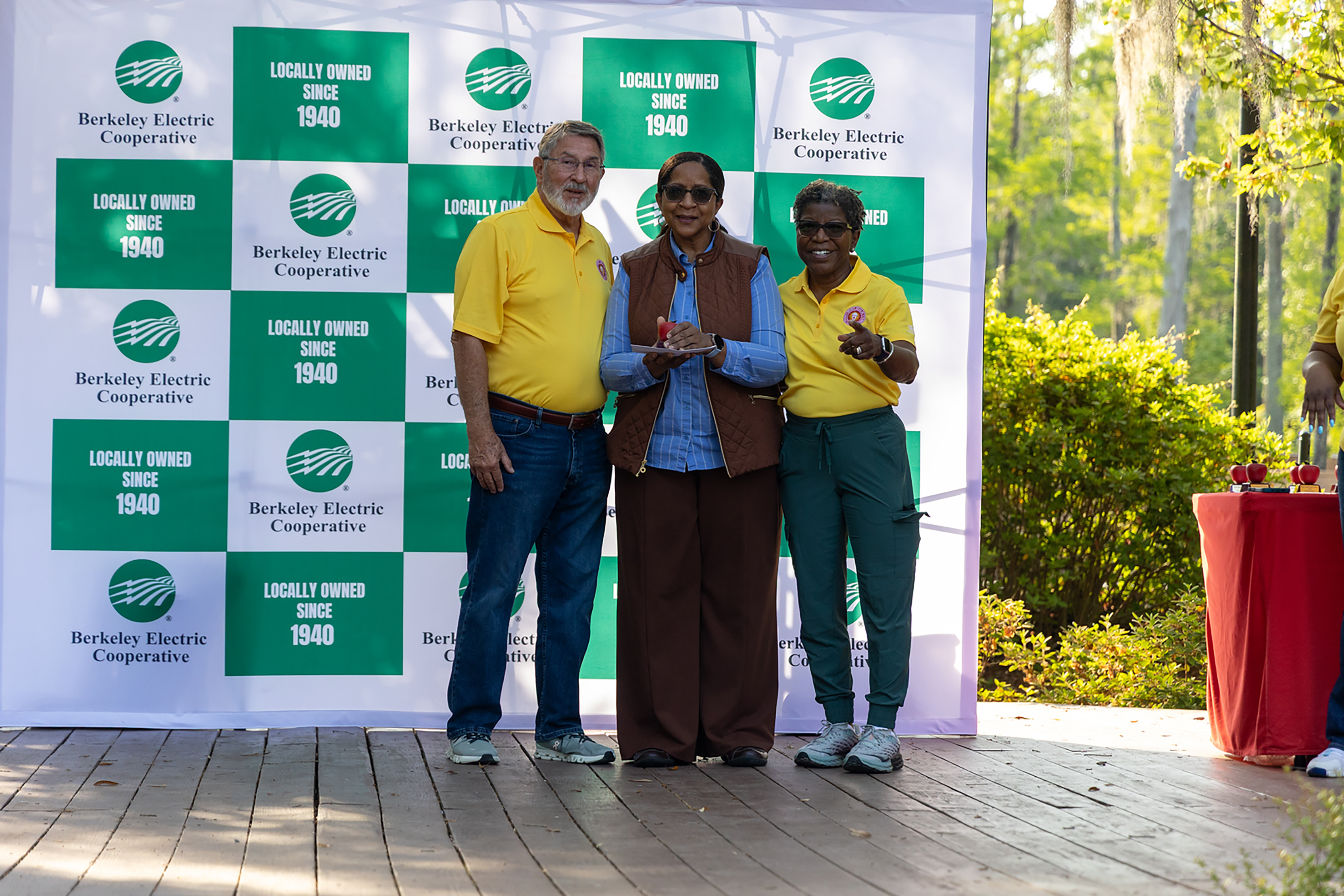 Three individuals stand on a wooden platform, smiling for a photo. A large banner with green, white, and red panels is behind them.