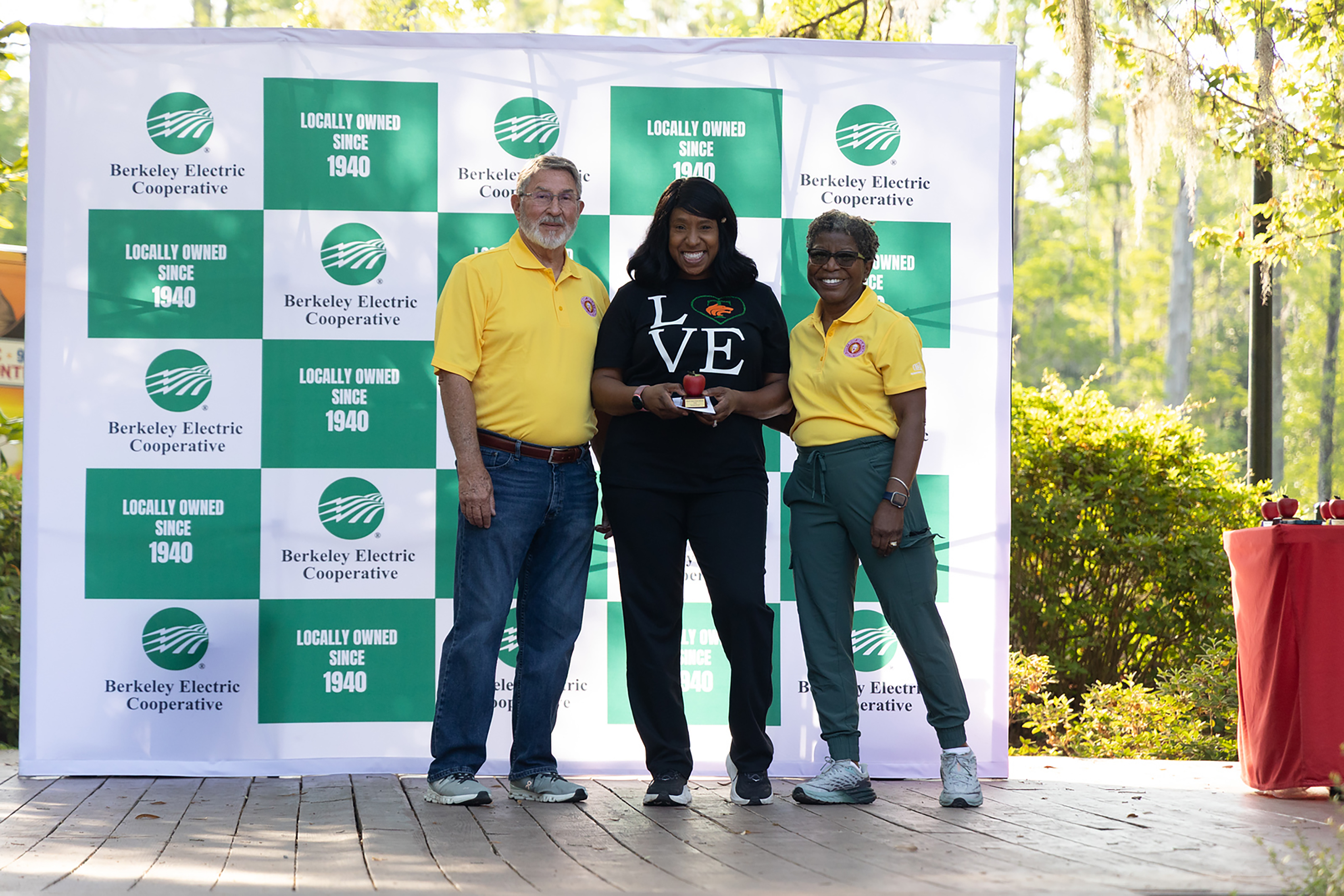 Three people in yellow shirts and one in green pants stand in front of a large green and white banner.