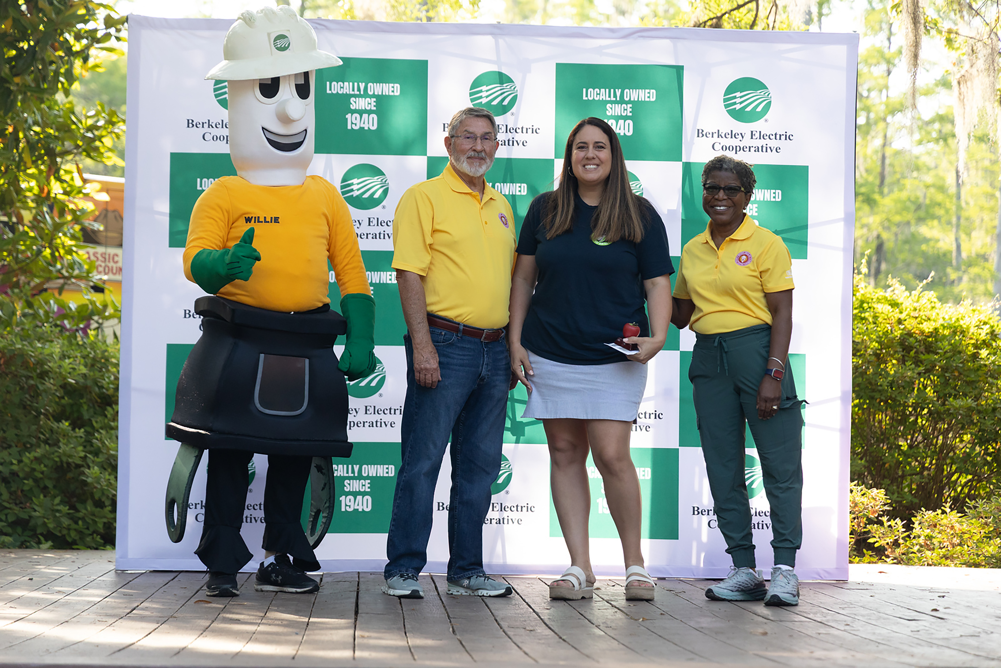 Four individuals on stage with a mascot; two men and two women wear yellow shirts. A woman holds a notebook.