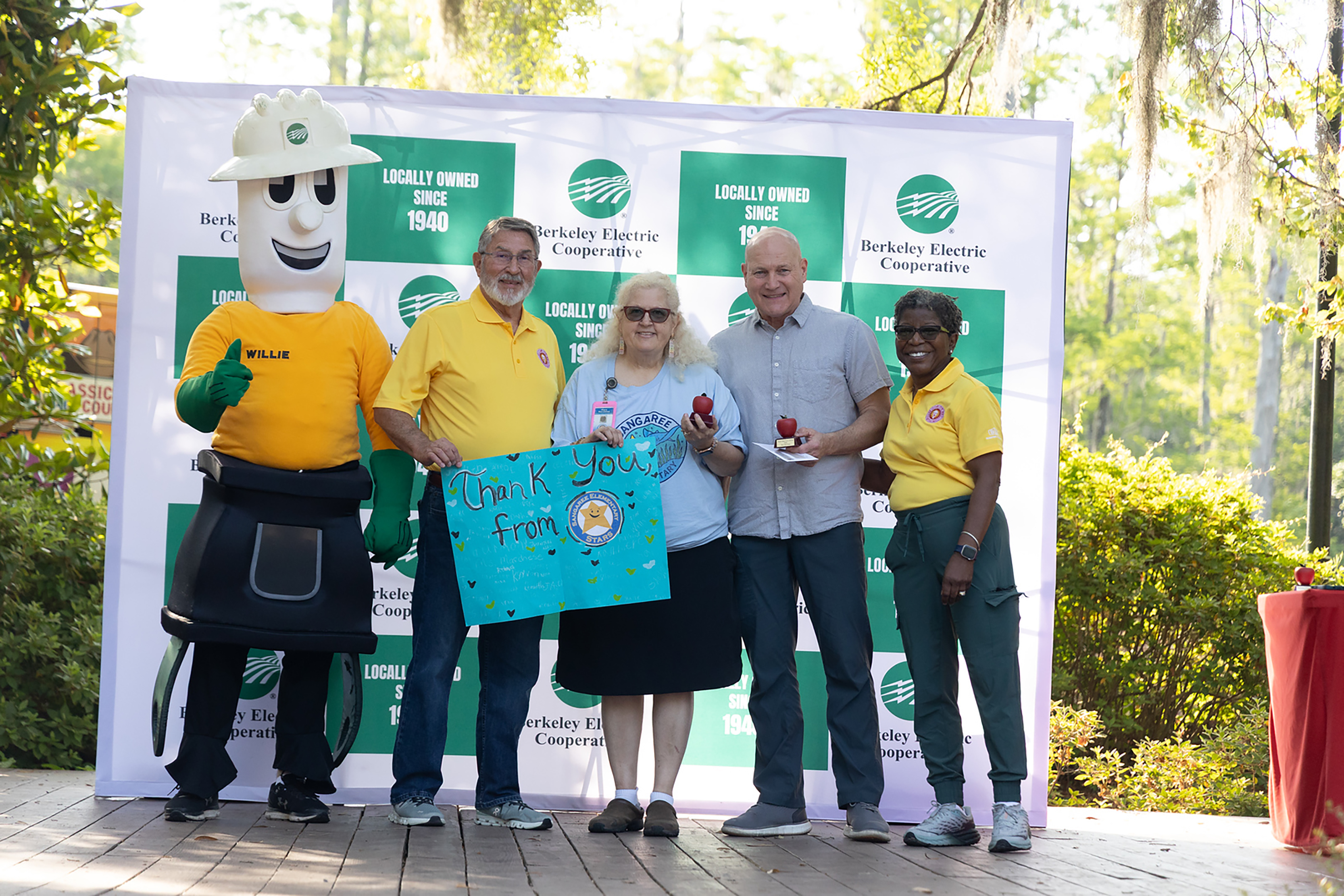 Five people, including a mascot, stand in front of a large banner with green and white sections.