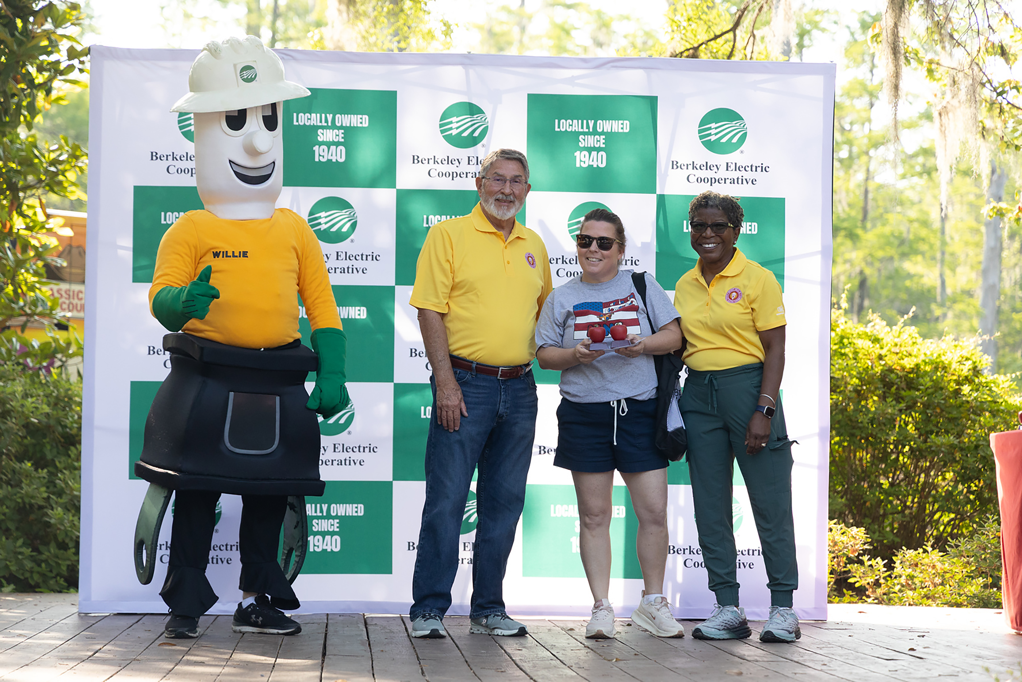 Four people stand in front of a green and white backdrop. The two women wear yellow shirts and pants. The man wears a yellow shirt and jeans.