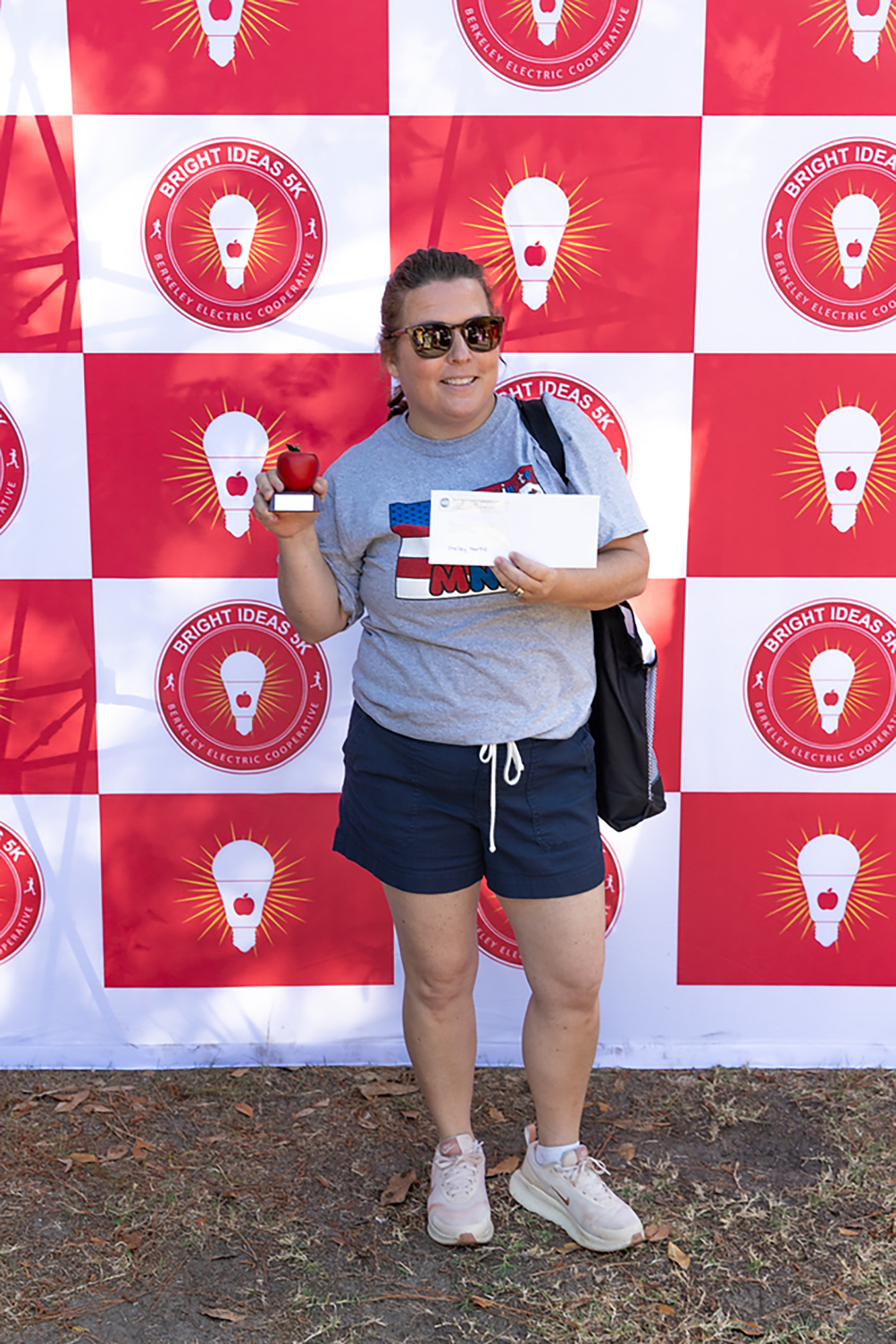Woman holding an apple and a certificate in front of a red and white checkered backdrop with light bulb logos.