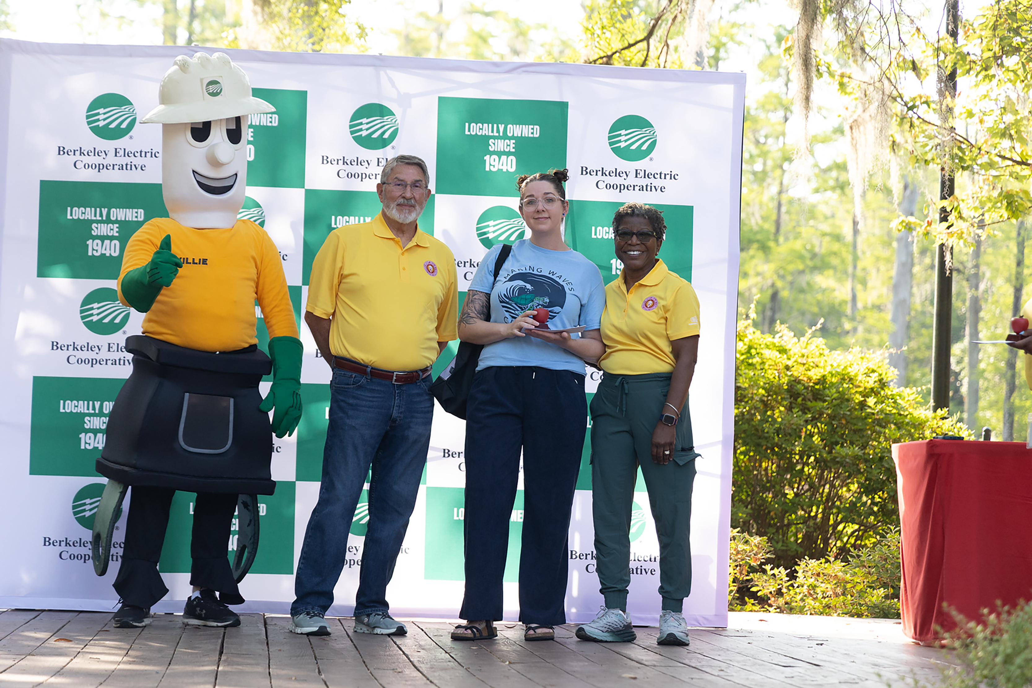 Four people pose on a stage with a mascot, all in yellow shirts. Green and white banners are behind them.