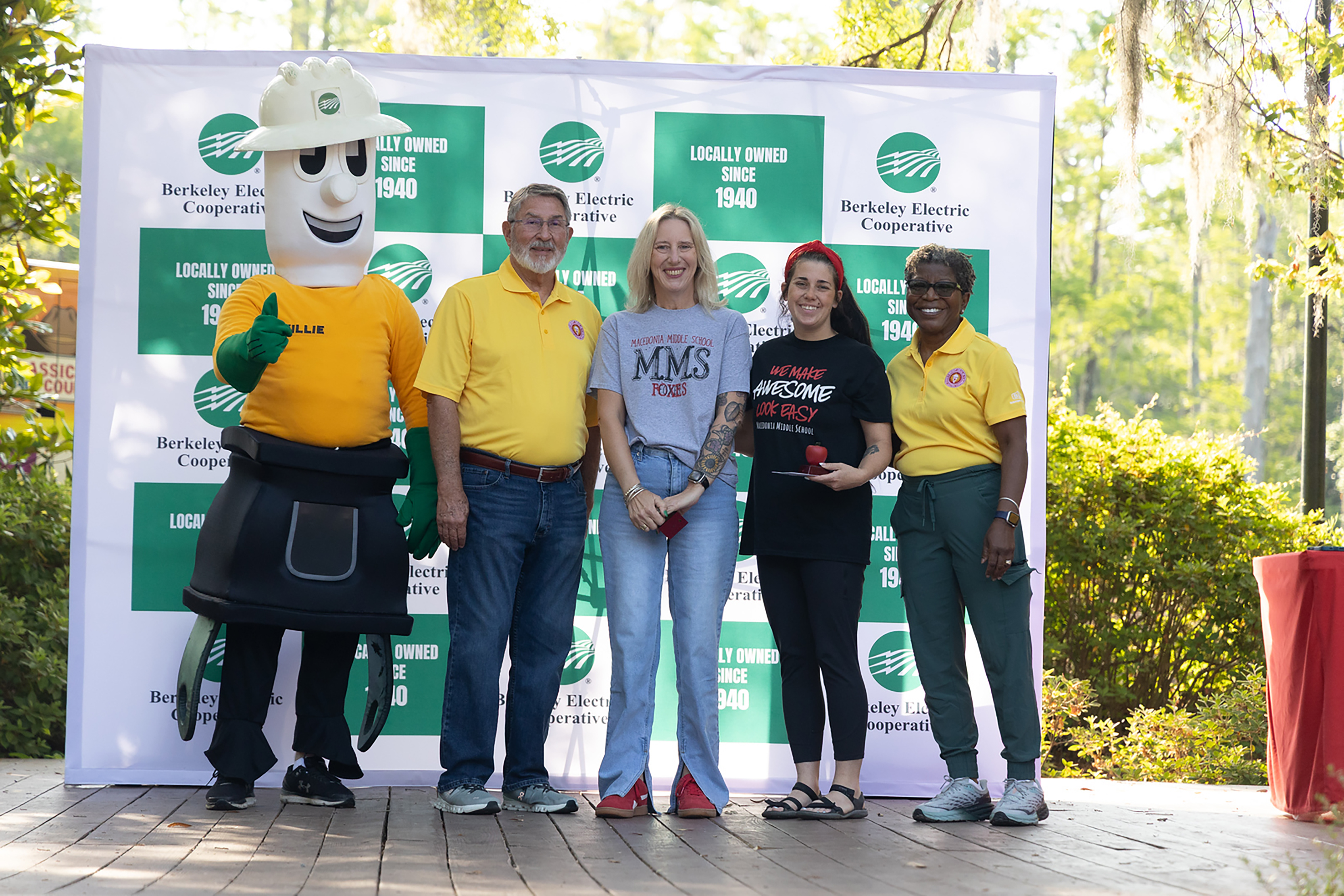Five people, including a mascot, stand in front of a backdrop with text and logos.