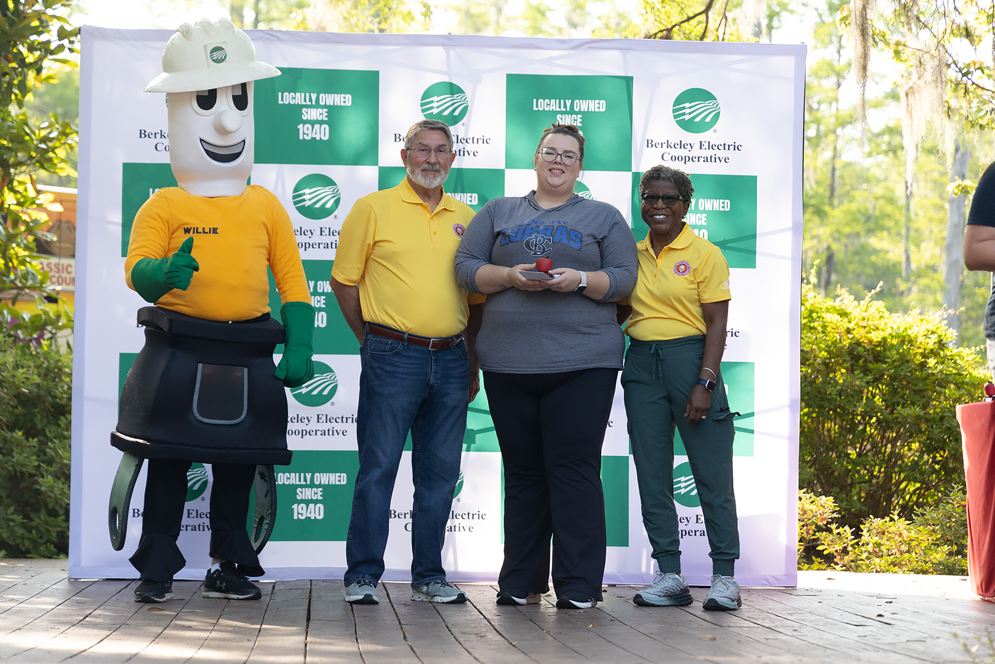 Four people in yellow and gray shirts stand by a mascot in front of a banner.