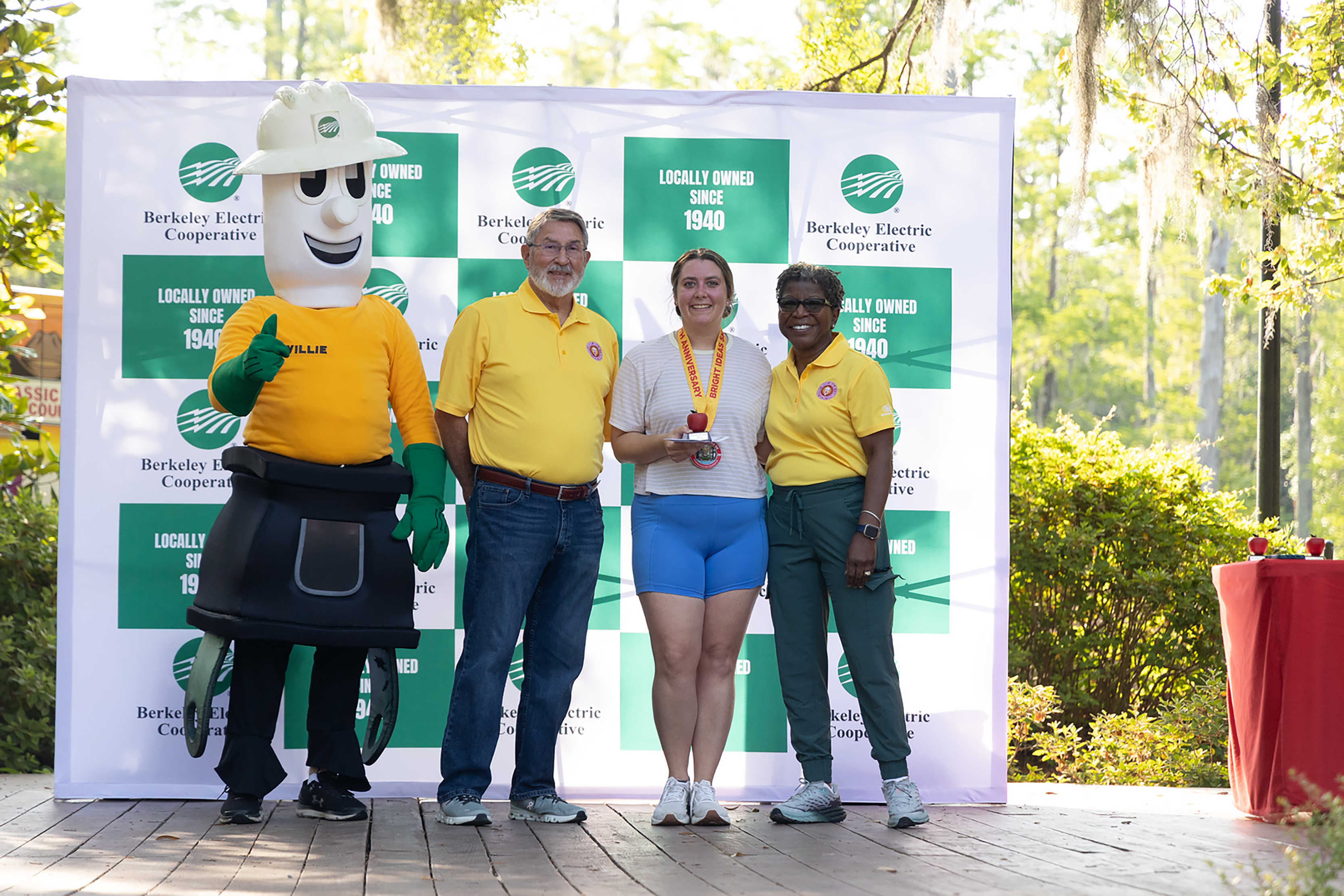 Four people stand behind a backdrop. Three of them wear yellow shirts, and one wears a mascot costume.