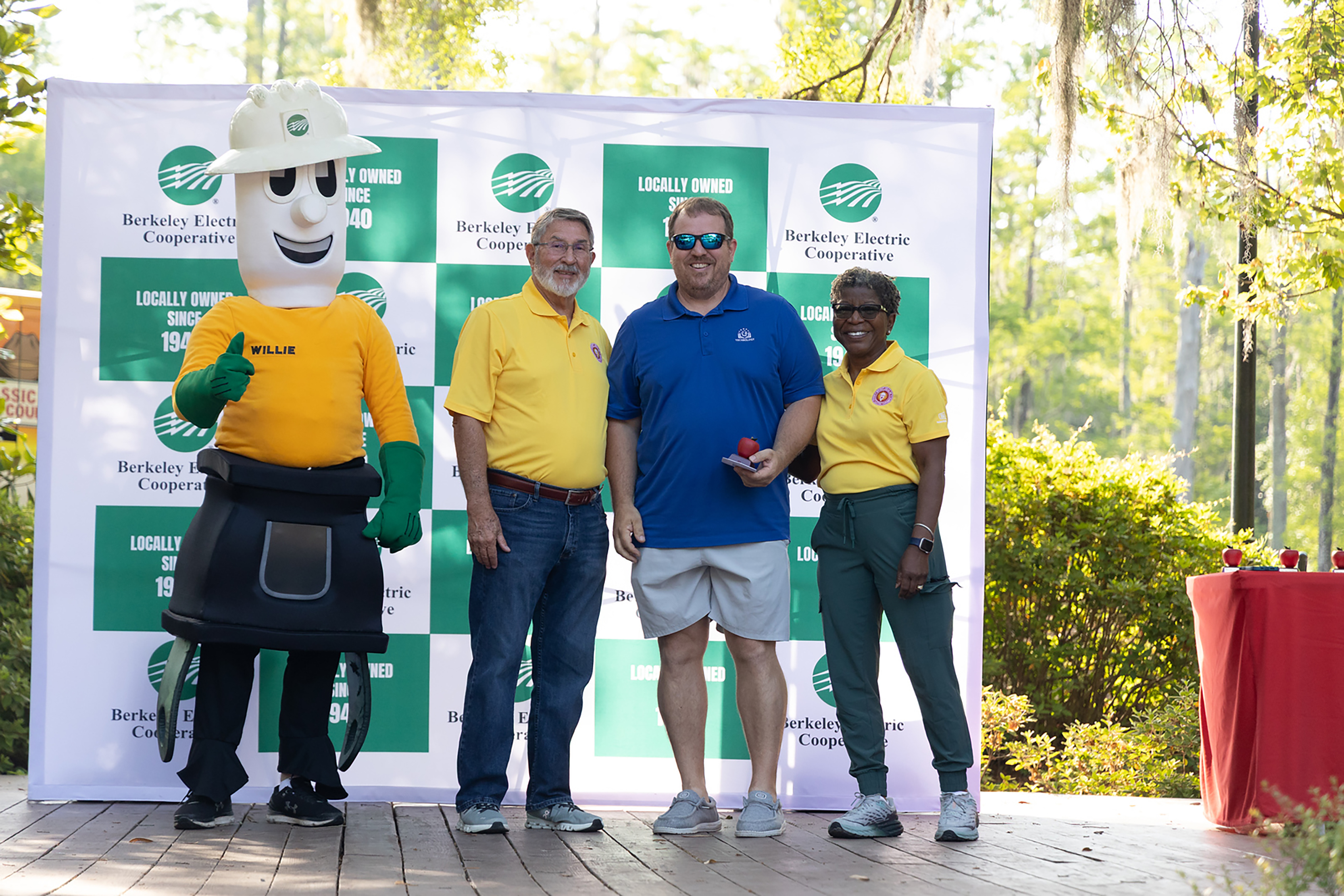 Three people stand on a wooden platform, one wearing a utility worker mascot costume, while two others wear yellow shirts.