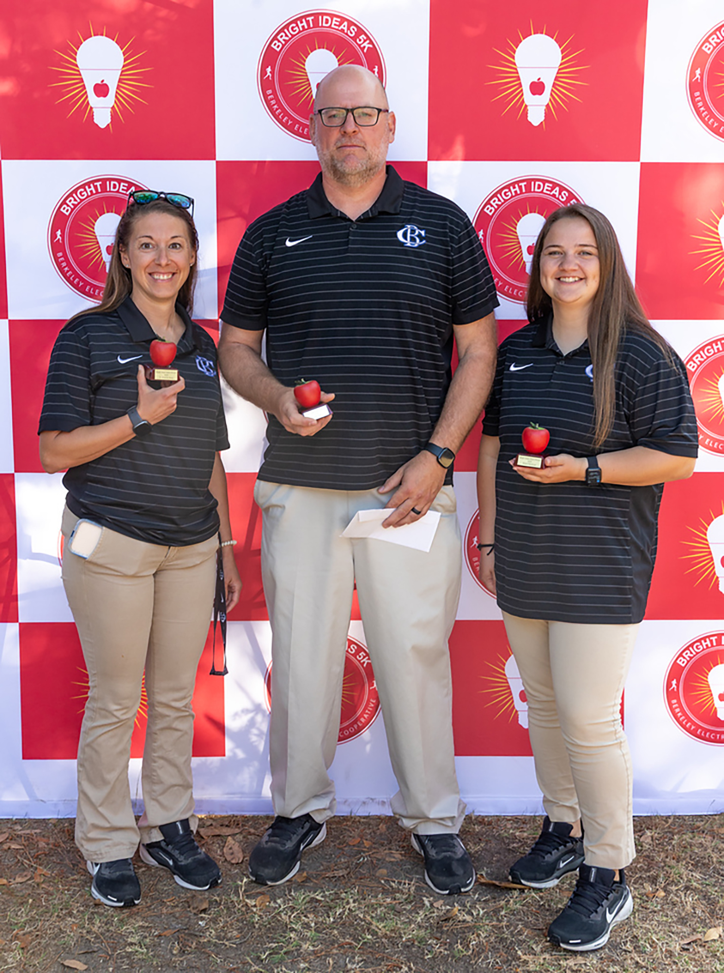 Three people in black and tan uniforms and glasses stand together. A man holds an apple and paper.