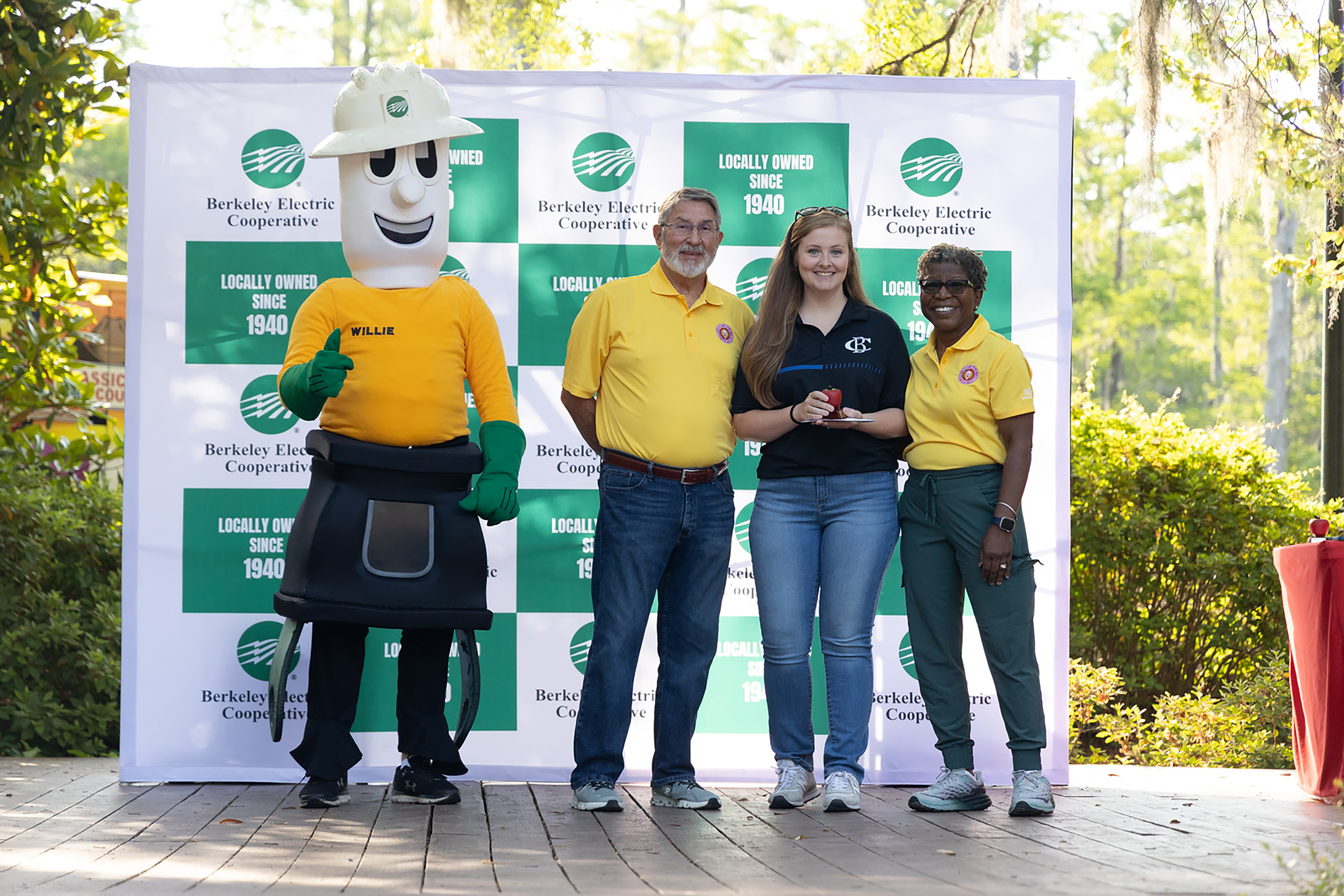 Four individuals, including a mascot, stand in front of a green banner, with three wearing yellow shirts.
