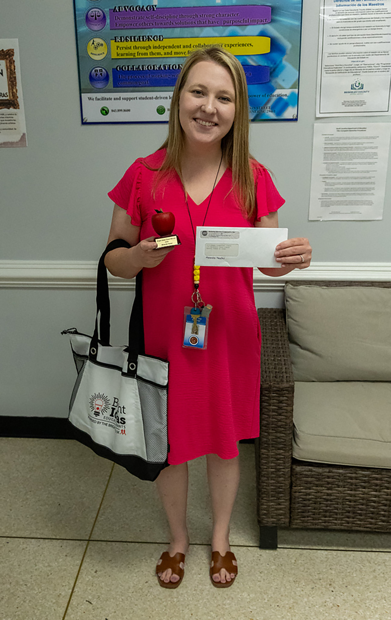 A woman in a pink dress stands holding a certificate and apple award in a room with a chair and bulletin board.