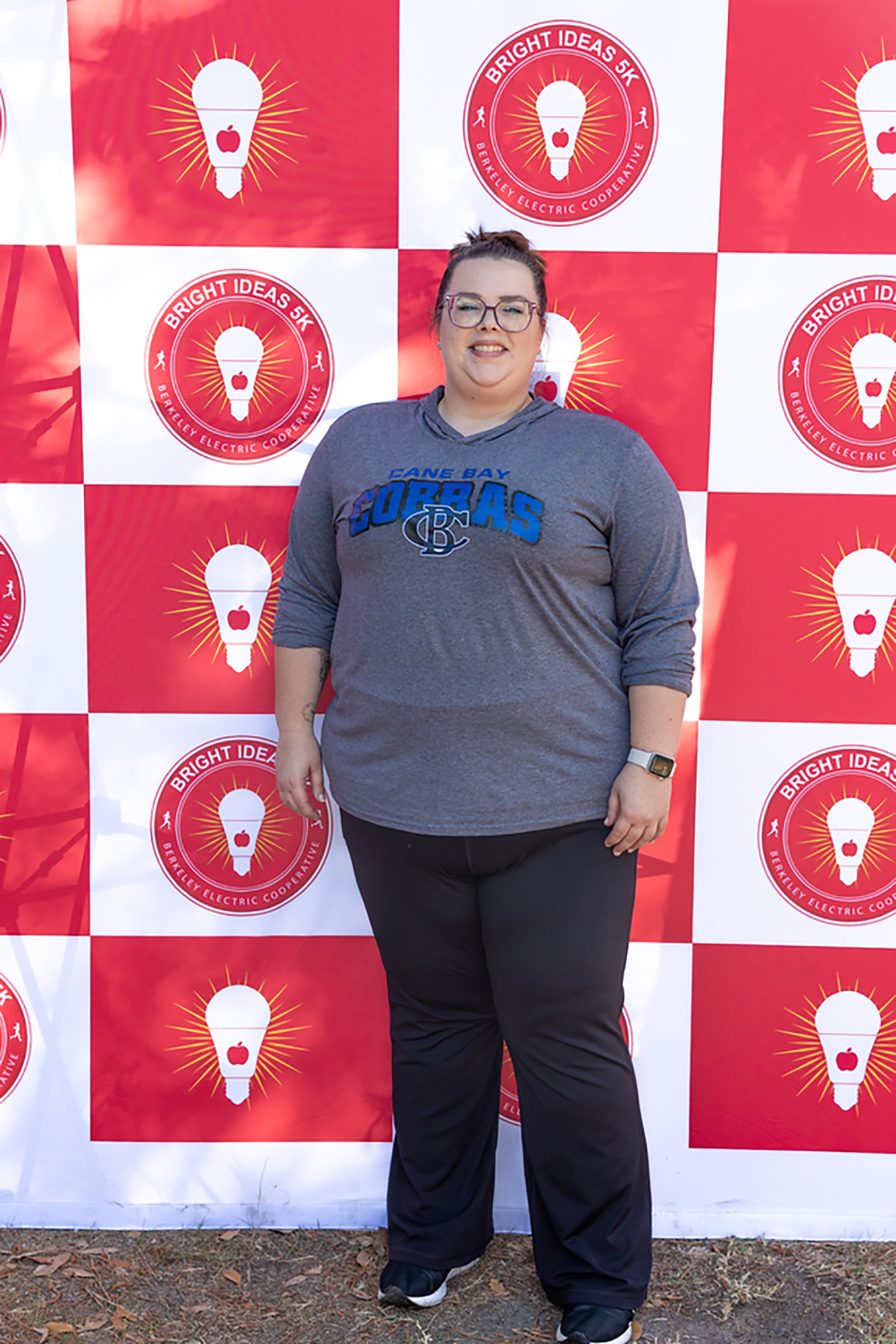A woman wearing a gray sweatshirt and glasses stands in front of a red and white checkered wall with light bulb logos.