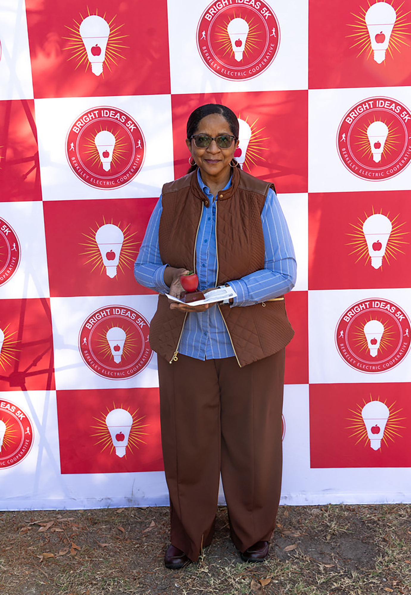 A woman in a blue shirt and brown vest stands before a red and white wall with light bulbs.
