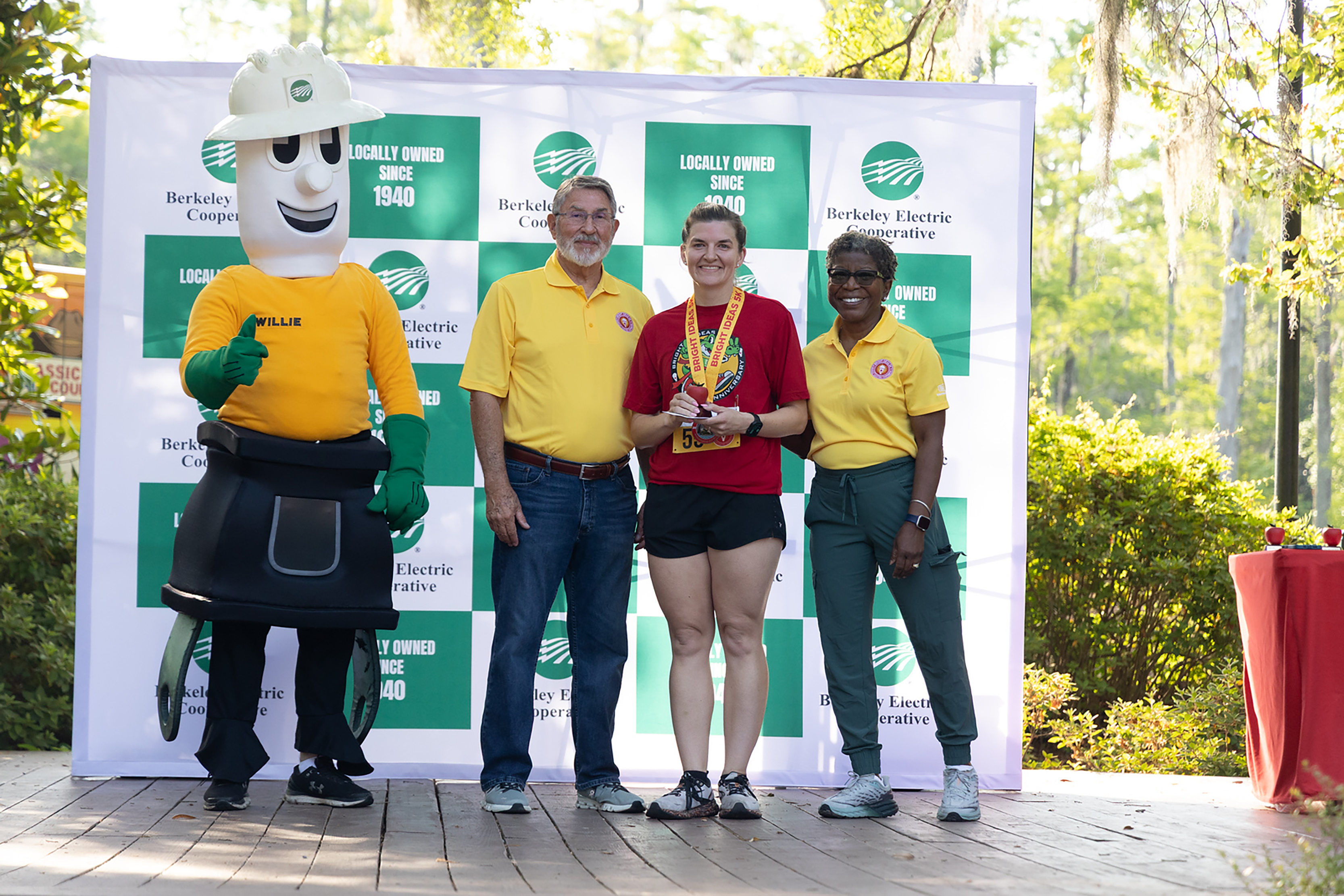 Four people stand together in front of a green and white banner with a mascot and plants behind them.
