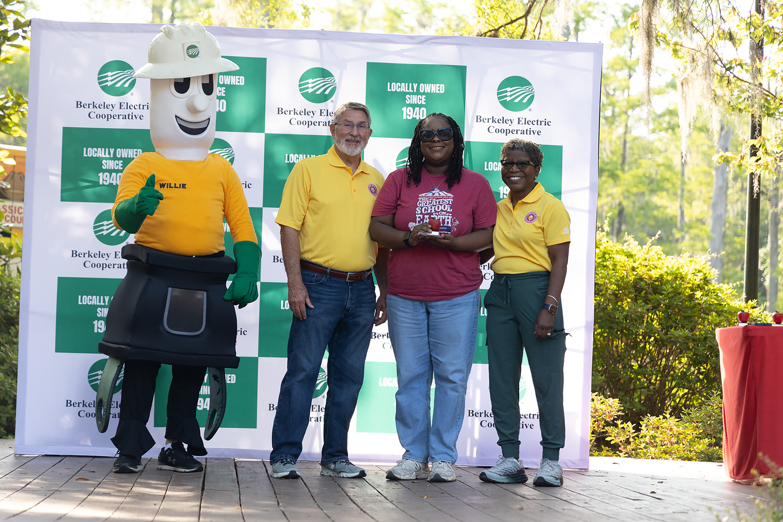 A man in a yellow shirt and three women in yellow and green attire stand in front of a banner.